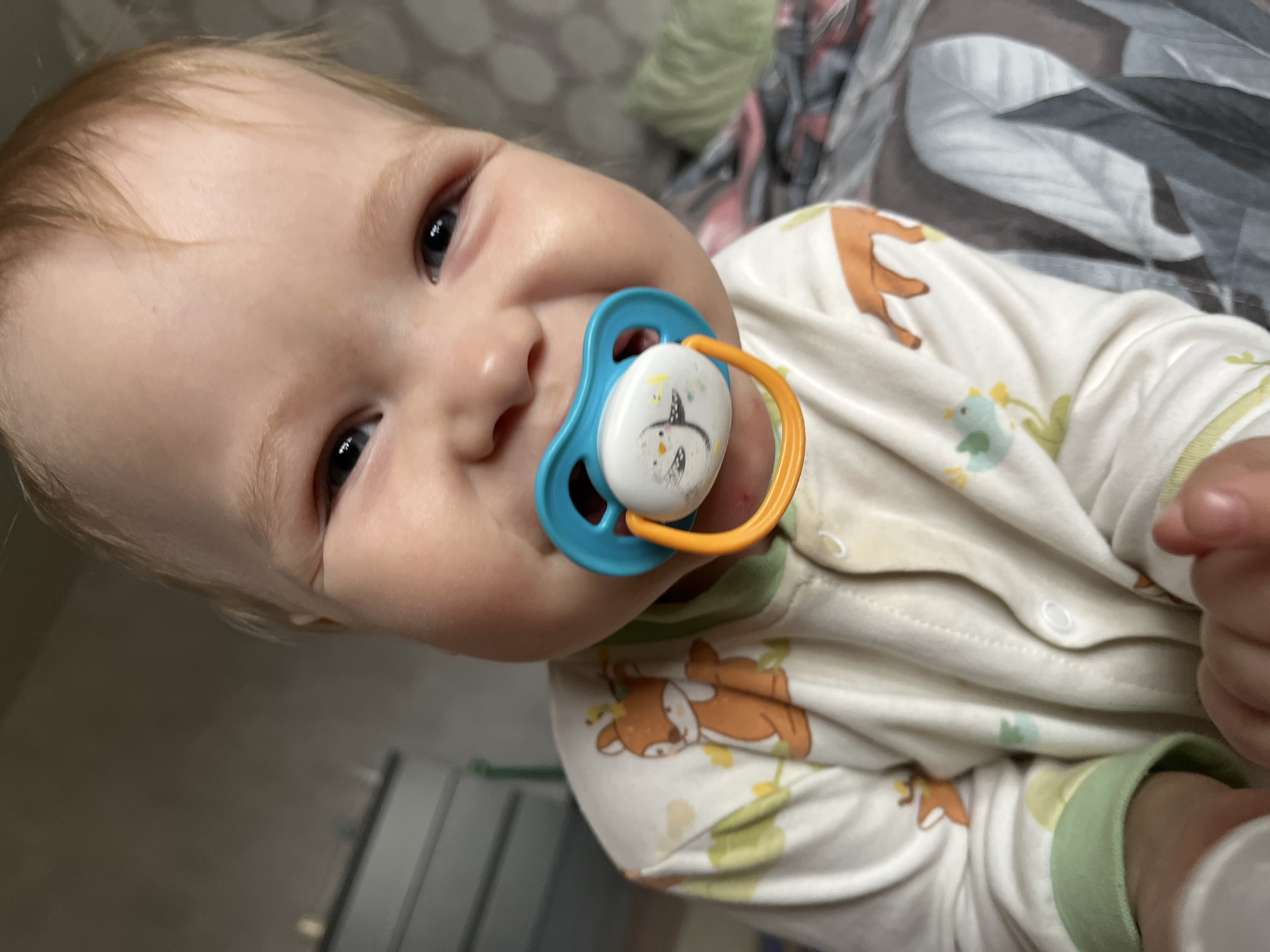 An infant or young toddler with light hair and blue eyes is depicted in a close-up, head-and-shoulders portrait. The infant is smiling broadly at the camera, with a pacifier in its mouth. The pacifier features a blue rim, an orange handle, and a white central disk adorned with a black and white bird graphic and yellow star-like elements. The child is wearing a light-colored, long-sleeved onesie or sleepsuit patterned with cartoon orange deer-like animals, light blue birds, and green and yellow foliage, with light green cuffs. Faint yellow staining is visible on the chest area of the garment. The background on the left shows a blurred light grey wall above a grey dresser or set of drawers. On the right, blurred dark and light patterned bedding, likely from a bed, is visible, along with a light green textured fabric item, possibly a pillow or blanket, on the far right. The scene is set indoors, within a residential environment, in the city of Chaikovsky, Russia.
