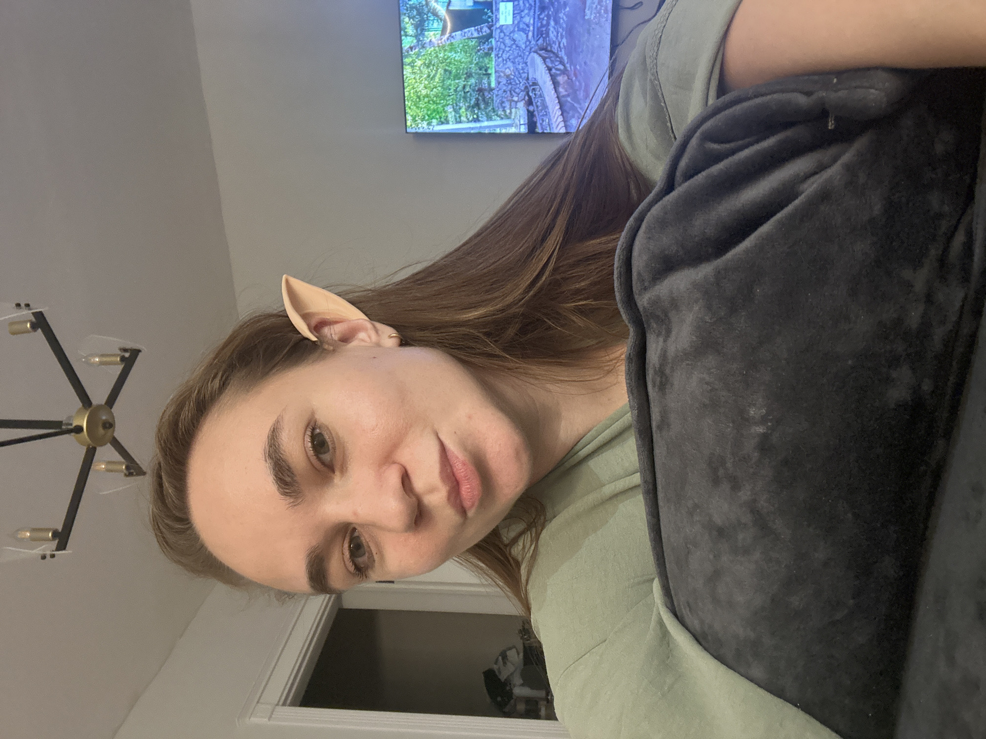 A woman is pictured indoors, presumably in a home or apartment, as indicated by the presence of a chandelier, a television displaying nature footage, and a doorway visible in the background. She is wearing what appears to be an elf ear prosthetic and is holding a dark grey, possibly velvet, pillow or cushion close to her. The woman is wearing a light green shirt. The location is not discernible as Kharnavy, Russia, from the visual information provided.