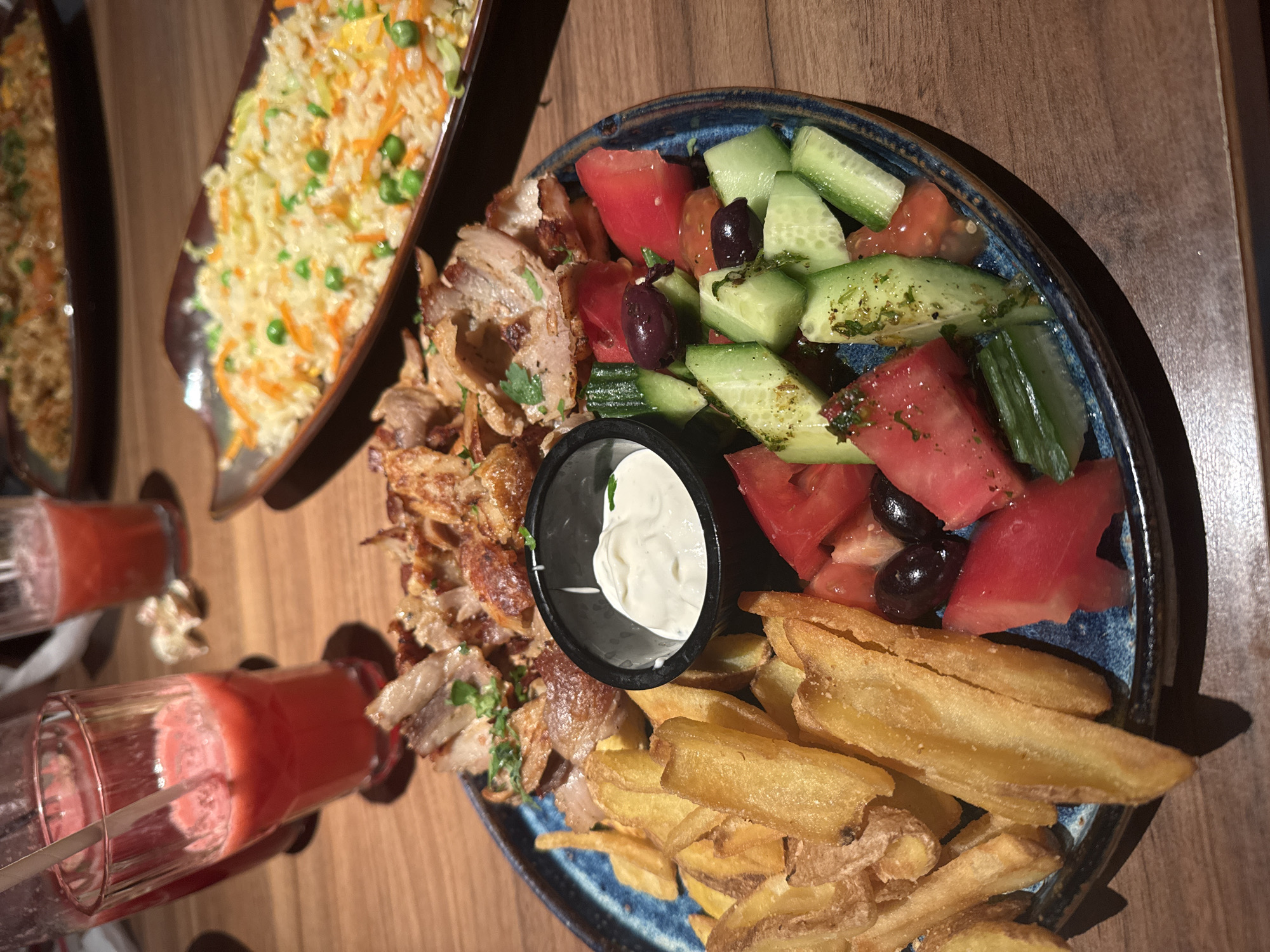 The image depicts a dining table laden with food and beverages in an indoor setting, likely a restaurant, in the city of Unknown, Unknown. The primary subject is a blue-rimmed round plate in the foreground, containing a mixed meal. This plate holds a portion of grilled or pan-fried sliced meat, possibly döner or gyros, garnished with green herbs. Adjacent to the meat is a generous serving of golden-brown, thick-cut french fries. A small black ramekin, centrally positioned on the plate, contains a white, creamy sauce. Completing the main plate is a fresh salad comprising diced red tomatoes, sliced green cucumbers, and several dark, whole olives.
In the mid-ground, to the right, two elongated, oval-shaped plates are visible, each containing a rice dish. The closer rice dish features white rice mixed with visible pieces of orange carrots and green peas, with lighter vegetable components also present. The further rice plate is similar but slightly out of focus.
In the background, to the left, multiple clear glasses are present, filled with a reddish-pink beverage, likely a fruit juice or smoothie; one glass contains a straw. The table surface is made of dark-grained wood. The lighting is warm and directional, creating shadows under the objects. No individuals are visible in the scene, and no active interactions are depicted.