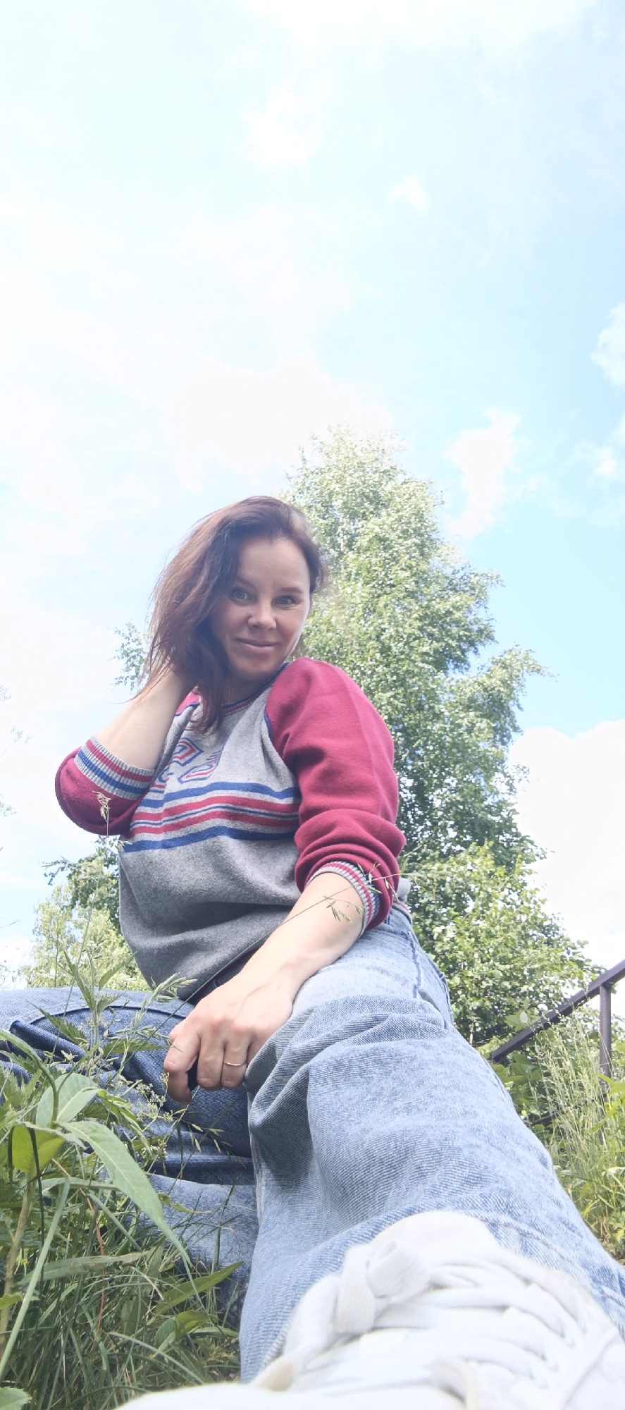 A low-angle, full-body shot captures a woman posing outdoors on a sunny day in Chaikovsky, Russia. The woman is sitting on what appears to be a step or raised platform, with her legs bent and crossed. She is wearing a grey sweatshirt with maroon sleeves, featuring horizontal blue and red stripes on the chest and striped cuffs. Light blue denim jeans and white sneakers complete her casual attire. Her right arm rests on her thigh, and her left hand is raised to her head, touching her hair. She is looking directly at the camera with a slight smile.
Behind her, a tall, leafy green tree dominates the background, with a bright blue sky dotted with white clouds above. The foreground is filled with lush green grass and vegetation, suggesting a natural or park-like setting. A dark railing is visible to the right of the frame, partially obscured by the foliage. The overall impression is of a relaxed, candid moment captured in an outdoor environment.