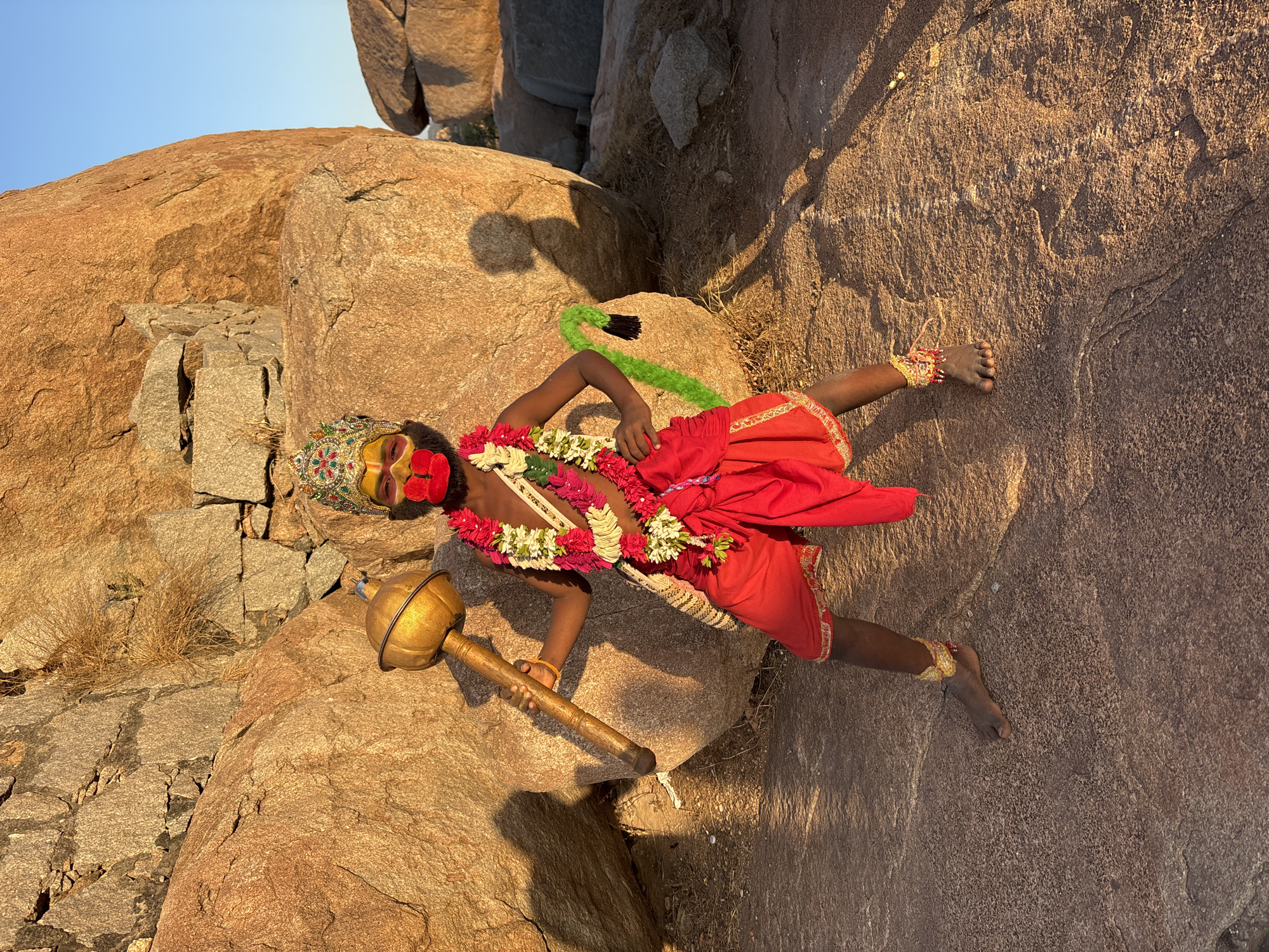 A young boy in a costume resembling the Hindu god Hanuman poses on a rocky terrain. He is dressed in red clothing, adorned with floral garlands and an ornamental headdress. His face is painted with yellow makeup and red around the mouth. He holds a golden mace in his left hand. A green and black faux tail is attached to his back. The scene is set against a backdrop of large reddish-brown boulders. The image was taken outdoors, with natural lighting. The location appears to be in the city of Kamalapura, India, given the characteristic rock formations.