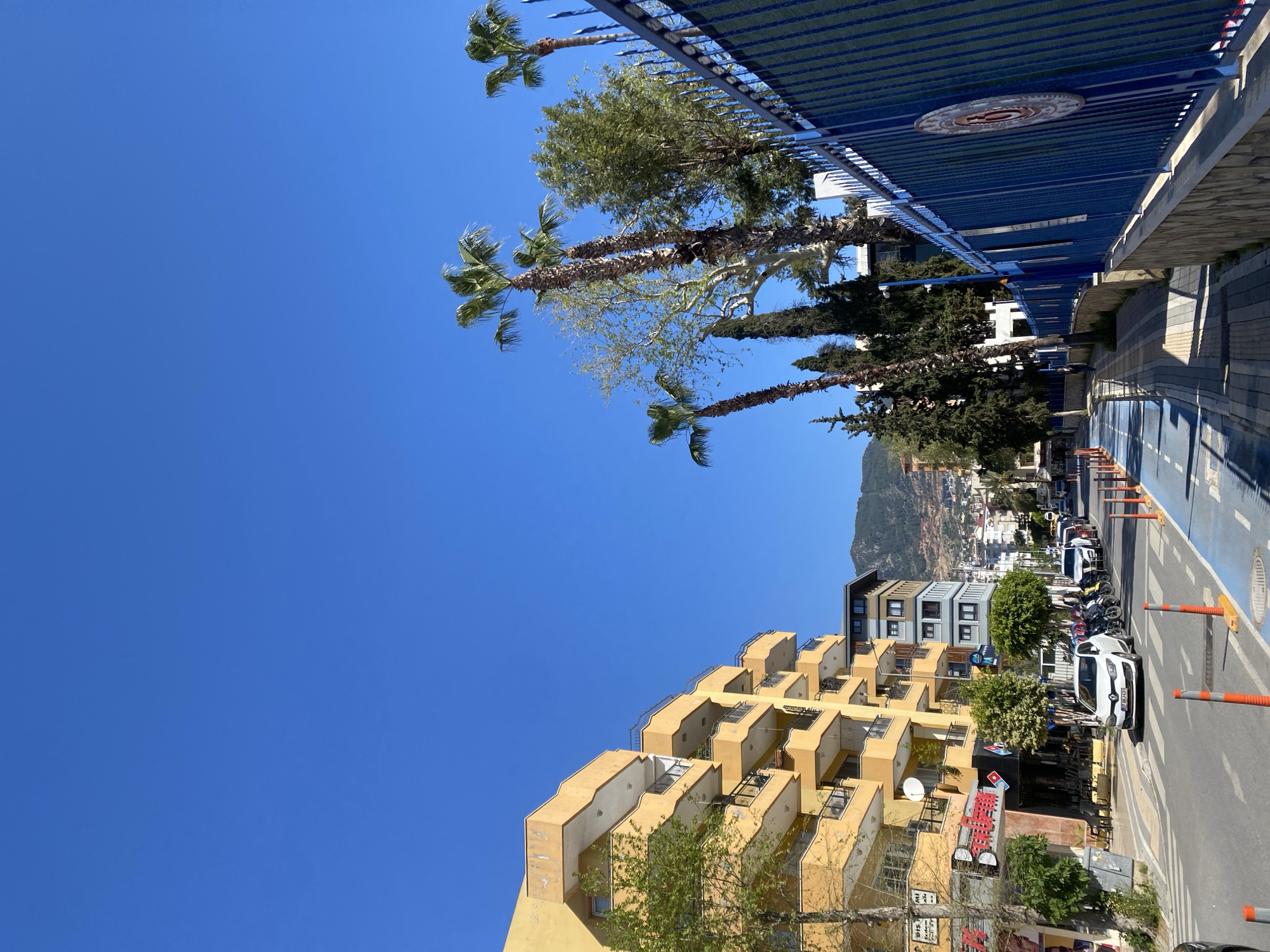 The image shows a street scene in Alanya, Turkey. The sky is clear blue. In the foreground is a yellow apartment building with multiple balconies. A palm tree and other trees line the street. Cars and motorcycles are parked along the side of the street. A blue bicycle lane is visible next to the sidewalk. A mountain is visible in the background. A blue metal fence runs along the upper part of the frame. The image appears to be taken from a slightly low angle.