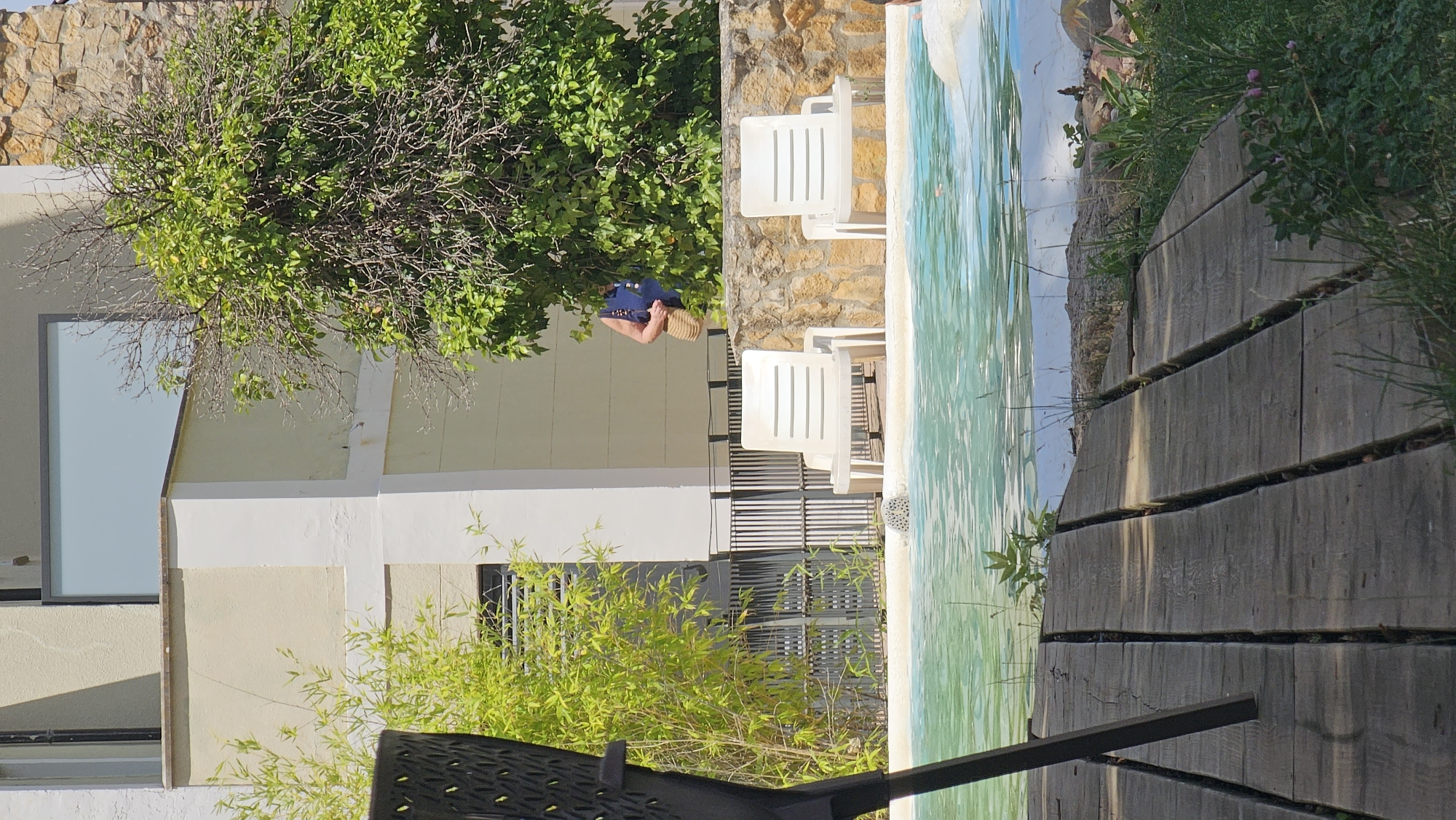 The image depicts an outdoor scene, likely a patio or poolside area, at Alhama De Aragón, Spain. A person, seen from the back, is leaning over and appears to be holding a blue bag and a straw hat or basket. They are positioned near a lush green tree with a stone wall in the background. To the right of the person, white plastic lounge chairs are set up next to a pool with a tiled mosaic bottom. The water in the pool has a bright turquoise hue. A wooden walkway made of planks runs along the edge of the pool, with grass and small plants growing in the gaps and along the sides. The scene is bathed in sunlight, suggesting it is daytime. The overall impression is one of relaxation and leisure in a warm climate.