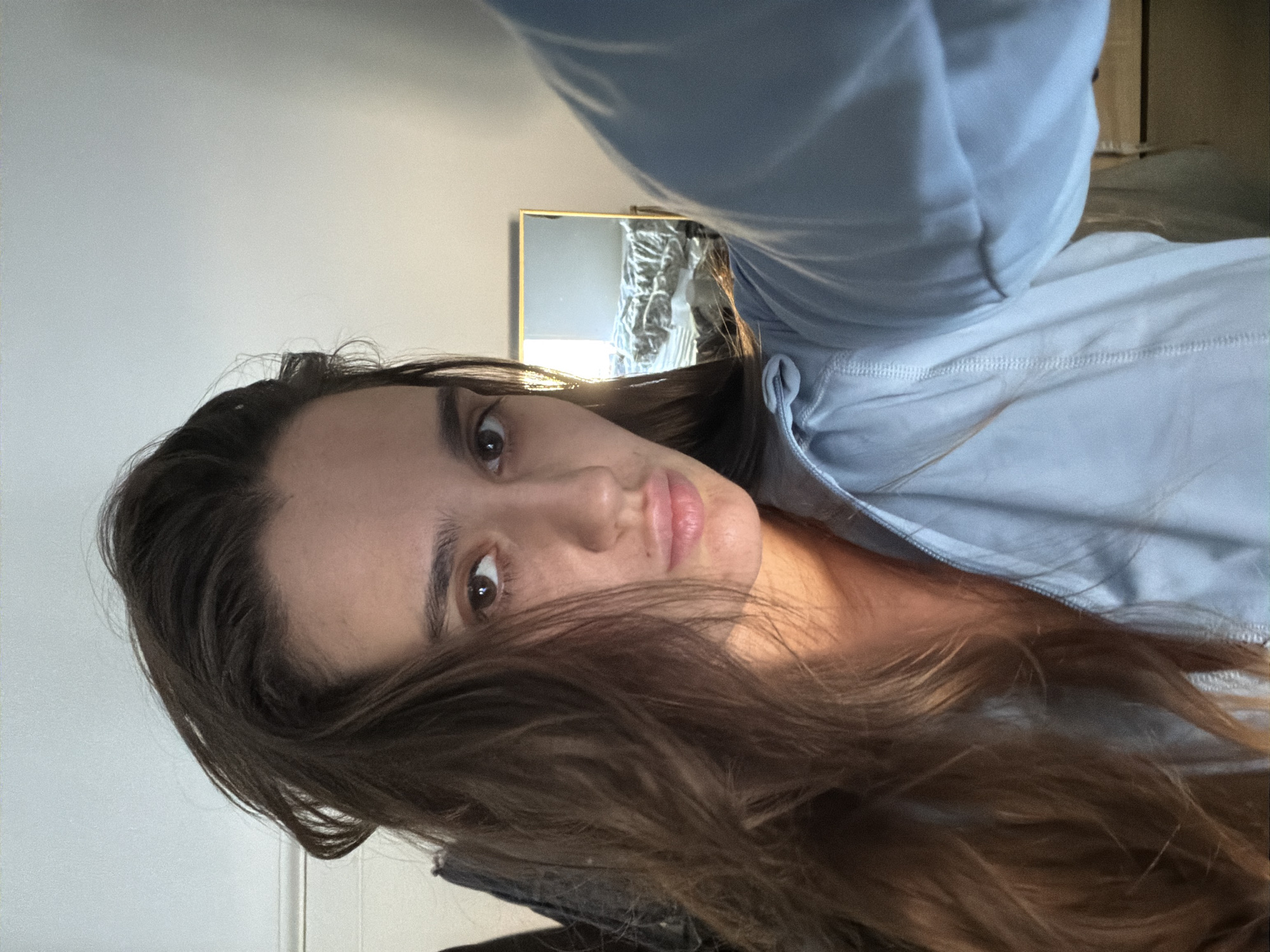 The image displays a close-up, selfie-style portrait of a single individual, a young adult female, situated indoors. She possesses long, dark brown, wavy hair, which partially frames her face and extends over her shoulders. Her eyes are brown, and her lips are slightly pursed in a neutral expression. She is dressed in a light blue, long-sleeved top or jacket with a visible zipper, partially unzipped at the collar.
Natural light, originating from the left side of the frame (the subject's left), strongly illuminates her face and hair, creating distinct highlights and shadows. The immediate background features a light-colored wall or ceiling above and to the right, and darker, indistinct objects, possibly garments on a hanger, to the left. Over her right shoulder, a rectangular mirror with a thin gold-colored frame reflects a room. Within the reflection, an unmade bed with light-colored linens and a headboard is visible, along with a window to the left of the bed, indicating a separate source of bright natural light. The reflected environment suggests a casual, personal indoor space, possibly a bedroom.