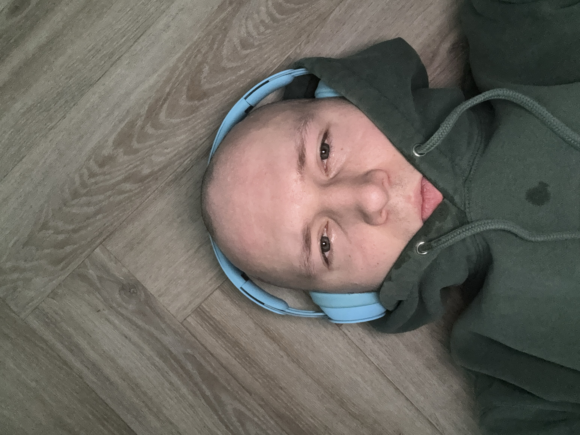 A bald person wearing light blue headphones and a green hooded sweatshirt lies on a wooden floor. The person's face is visible, and they are looking directly at the camera. The location in Krasnoyarsk, Russia, cannot be determined from the image.