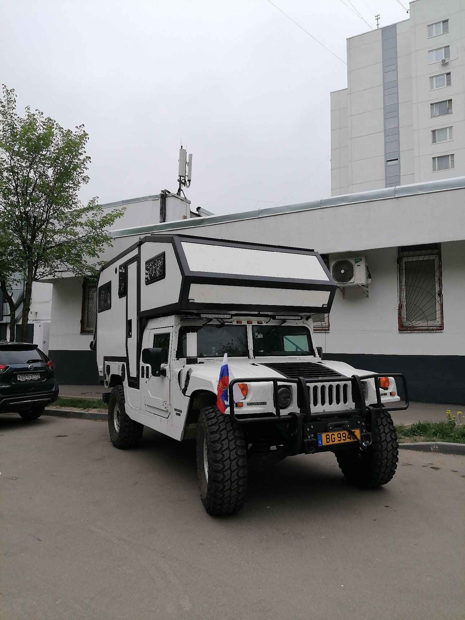 The image shows a customized white Hummer H1 parked on a street. The Hummer has a camper module attached to the chassis. The vehicle has a license plate that reads "BG 994", with a Serbian flag on the lower left. The vehicle appears to be parked in a residential area of Moscow, Russia. In the background, there is a tall white building with multiple windows and a grey accent running vertically. To the left of the Hummer is a dark-colored SUV. The overall lighting suggests an overcast day.