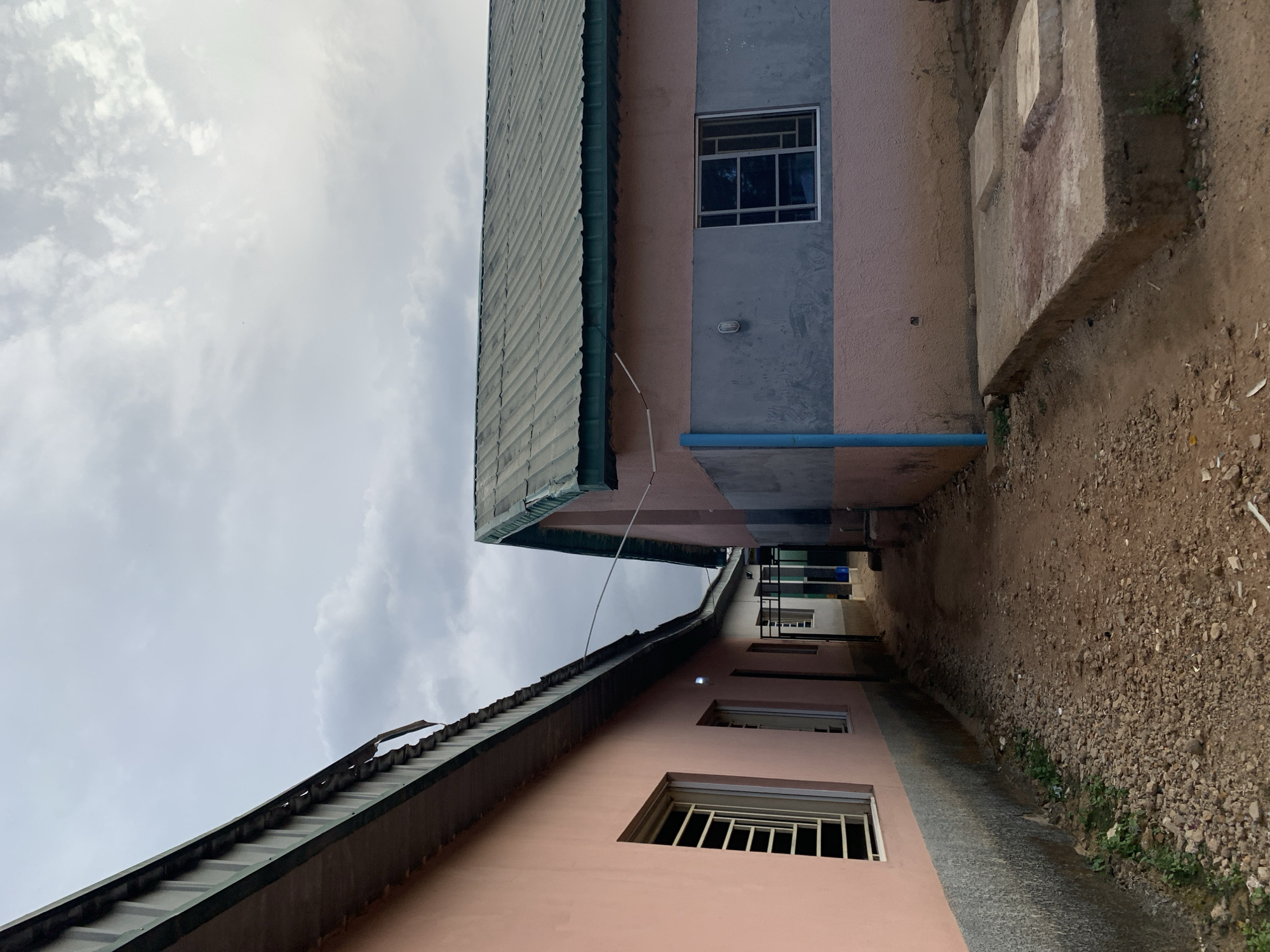 This image captures a narrow passage between two buildings, likely in New Stadium, Nigeria. The buildings are painted in shades of pink and grey with metal roofs and barred windows. A blue pipe runs horizontally across one of the building's exterior walls. The ground is a mix of dirt, small rocks, and sparse vegetation. The sky is overcast with grey clouds. The perspective is looking upwards along the alleyway, suggesting a ground-level viewpoint. The scene appears deserted with no visible people or signs of recent activity.