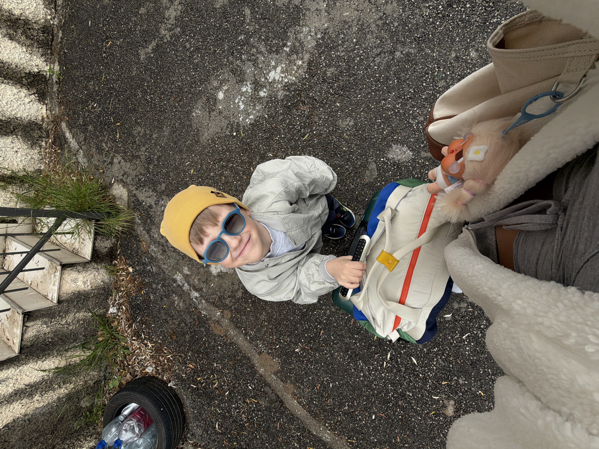 An overhead shot captures a young child smiling up at the camera, situated outdoors in Nerin, Montenegro. The scene is dominated by dark asphalt ground, showing patches of white residue and scattered dry leaves. To the left, concrete steps with a black metal railing ascend, bordered by sparse green grass.
The primary subject is a fair-haired boy, appearing to be a toddler or preschooler. He wears a bright yellow beanie, stylish blue-rimmed sunglasses, a light grey zippered jacket, and dark sneakers. He is holding a black and white electronic-looking device in his right hand. The child's gaze is directed upwards towards the camera, and he has a clear, happy smile on his face.
Partially visible on the right side of the frame is an adult, likely holding the camera, indicated by a leg in grey trousers, an arm or body covered by a cream-colored fluffy garment, and a beige canvas bag with brown accents. Attached to this bag are a blue plastic clip and a small, light-haired doll with a flower on its head, resembling a Troll doll.
Next to the child, a beige and colorful backpack or a part of a stroller with red zippers, a yellow strap, and blue and green sections is visible. In the lower left corner, an old black car tire lies on its side, serving as an impromptu container for several plastic water bottles, some clear and some red.
The time of day appears to be daytime, with diffused light suggesting either an overcast sky or indirect sunlight. No discernible text is visible on any objects. The overall impression is a casual, everyday outdoor moment captured from a unique perspective.