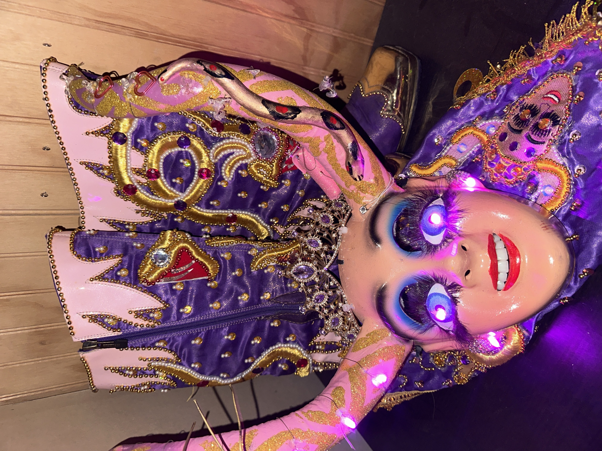 A brightly lit doll, possibly part of a costume, sits indoors against a wood-paneled wall, likely in Arica, Chile. The doll has a painted face with large, bright blue eyes, long eyelashes, and a wide red smile revealing white teeth. Its skin is a pale pink, and it's adorned with golden glitter designs. The doll is wearing a purple and pink costume heavily embellished with gold embroidery, pearls, and gemstones. The costume includes snake-like features painted on the arms, and other intricate designs on the bodice. Some elements are lit with purple lights. A metallic gold boot is partially visible. The overall style of the doll and costume suggests a cultural performance or festival attire.