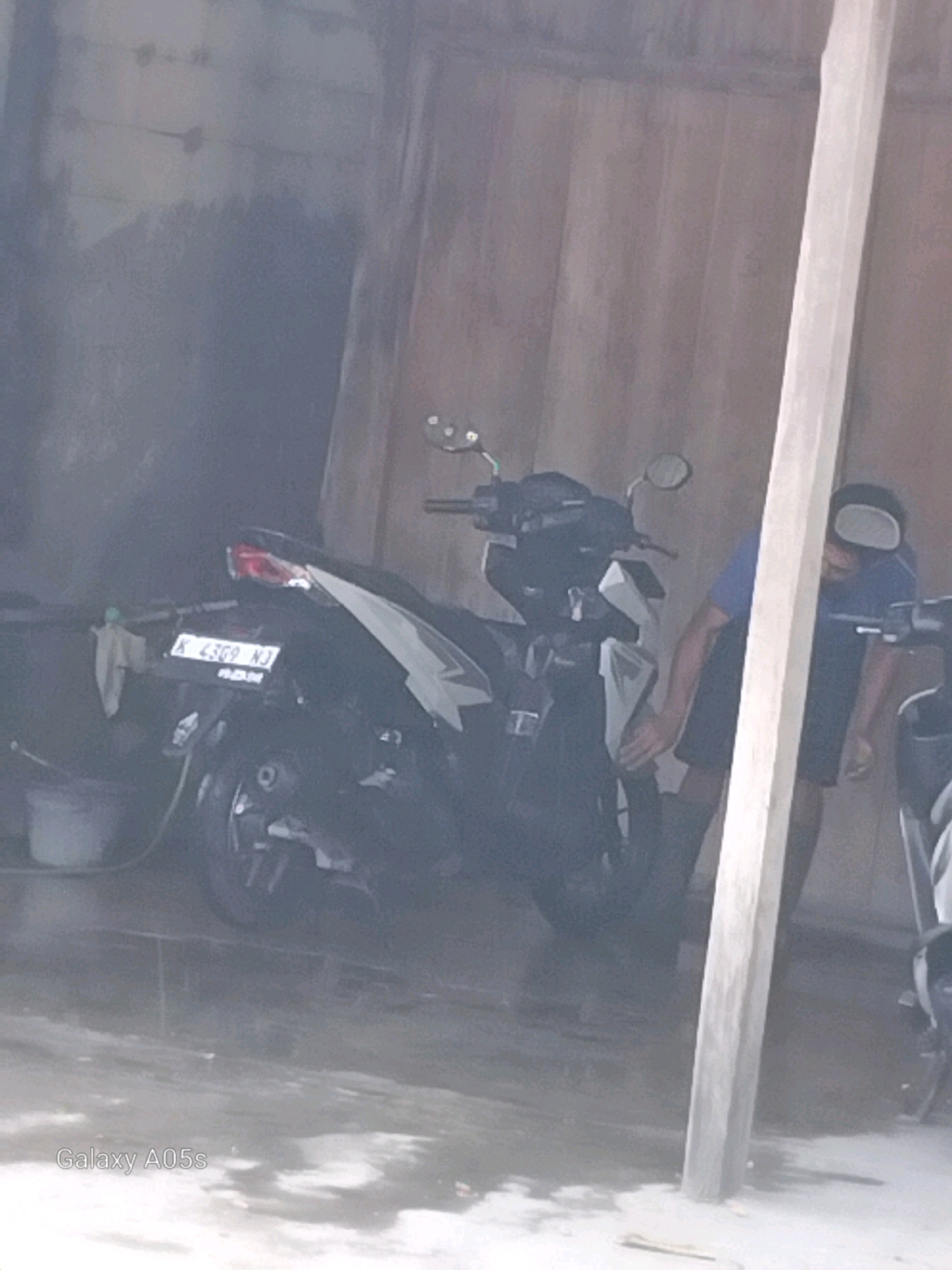 In a dimly lit, cluttered outdoor area, possibly a carport or a narrow alleyway, a person is seen standing behind a black motorcycle. The person, wearing a blue shirt and dark shorts, is partially obscured by a wooden post and the motorcycle itself. They appear to be interacting with the motorcycle in some way, though the exact action is unclear due to the low image quality and focus. The motorcycle, a modern scooter-type, has a license plate reading "K 4369 NJ". A white cloth is hanging from the handlebars. The ground is wet and reflective, suggesting recent rain or cleaning. The background consists of a corrugated metal wall and a vertical wooden beam. The scene is consistent with an area in Karangayung, Indonesia, where such motorcycles are common and outdoor spaces are often used for various activities.