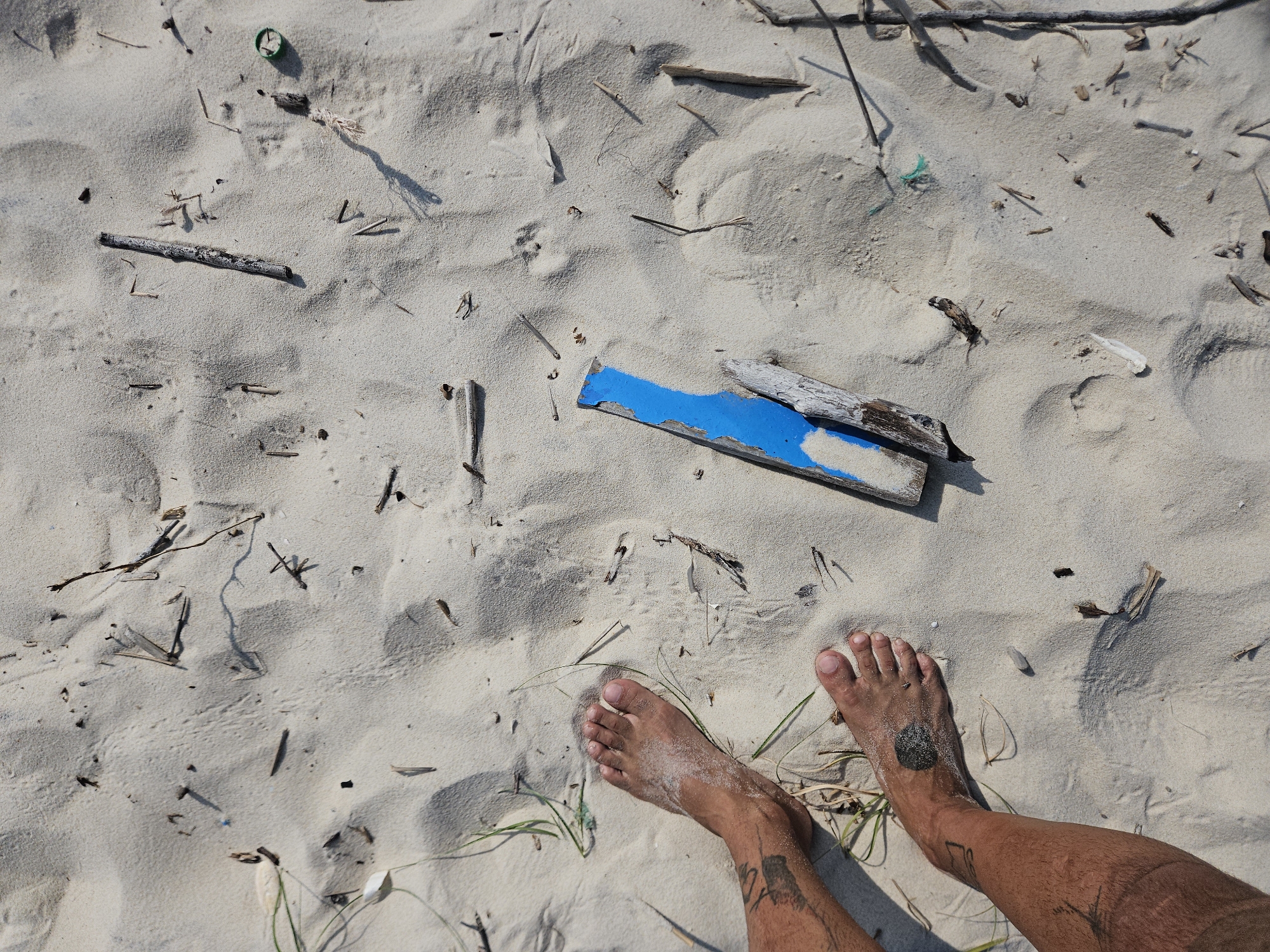 An overhead shot captures a person's bare feet standing on a sandy beach in Bac Cam Ranh Ward, Vietnam, under bright, clear sunlight. The scene is dominated by light, fine-grained sand, which shows subtle undulations and small footprints.
The person's two bare feet, visibly tanned and lightly covered in sand, are positioned in the lower right portion of the frame. The right foot features a distinct dark, circular tattoo on the top, and further up both lower legs, various linear and abstract tattoo designs are visible. Strong, sharp shadows cast by the feet, debris, and sand mounds indicate a sunny day, likely around midday or early afternoon.
The sand is littered with a variety of natural and man-made objects. Scattered throughout are numerous pieces of driftwood and small twigs. A prominent feature is a piece of wood, partially painted a vibrant blue, lying horizontally in the center. Other debris includes a green plastic bottle cap, fragments of white plastic or shell, a small piece of white rope, and some dried grass or weeds emerging from the sand. The presence of varied debris suggests a natural, perhaps less frequently cleaned, coastal environment. There is no discernible text visible on any objects or within the scene.