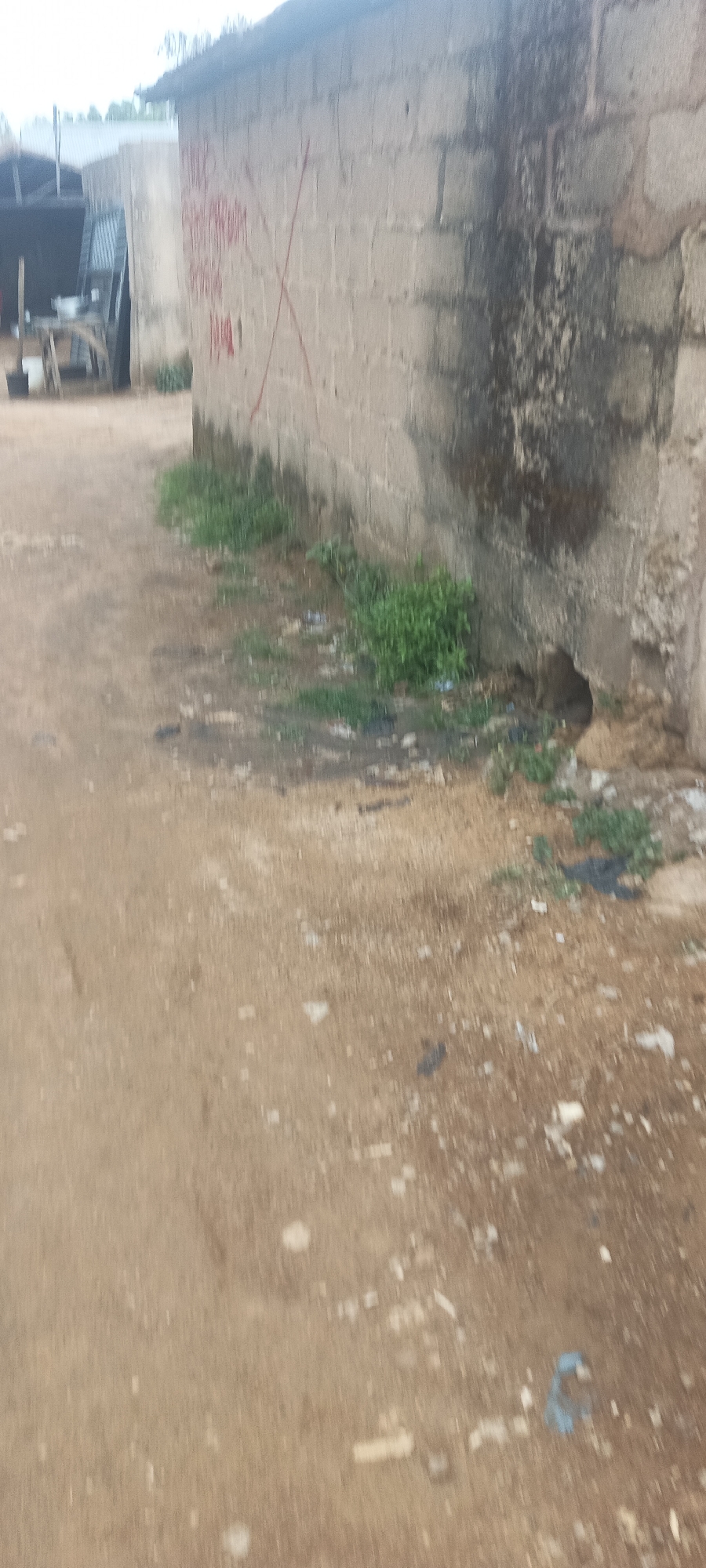 The image depicts a dirt road in Jos, Nigeria, adjacent to a concrete block building. The building's wall is marked with faded red graffiti and an "X" symbol. The base of the wall is discolored with moisture and has a small hole near the ground. Weeds and trash are scattered along the base of the wall and on the dirt road. In the background, there is a partial view of another building with a corrugated metal roof and some objects that appear to be buckets and other containers. The overall scene appears unkempt and possibly neglected.