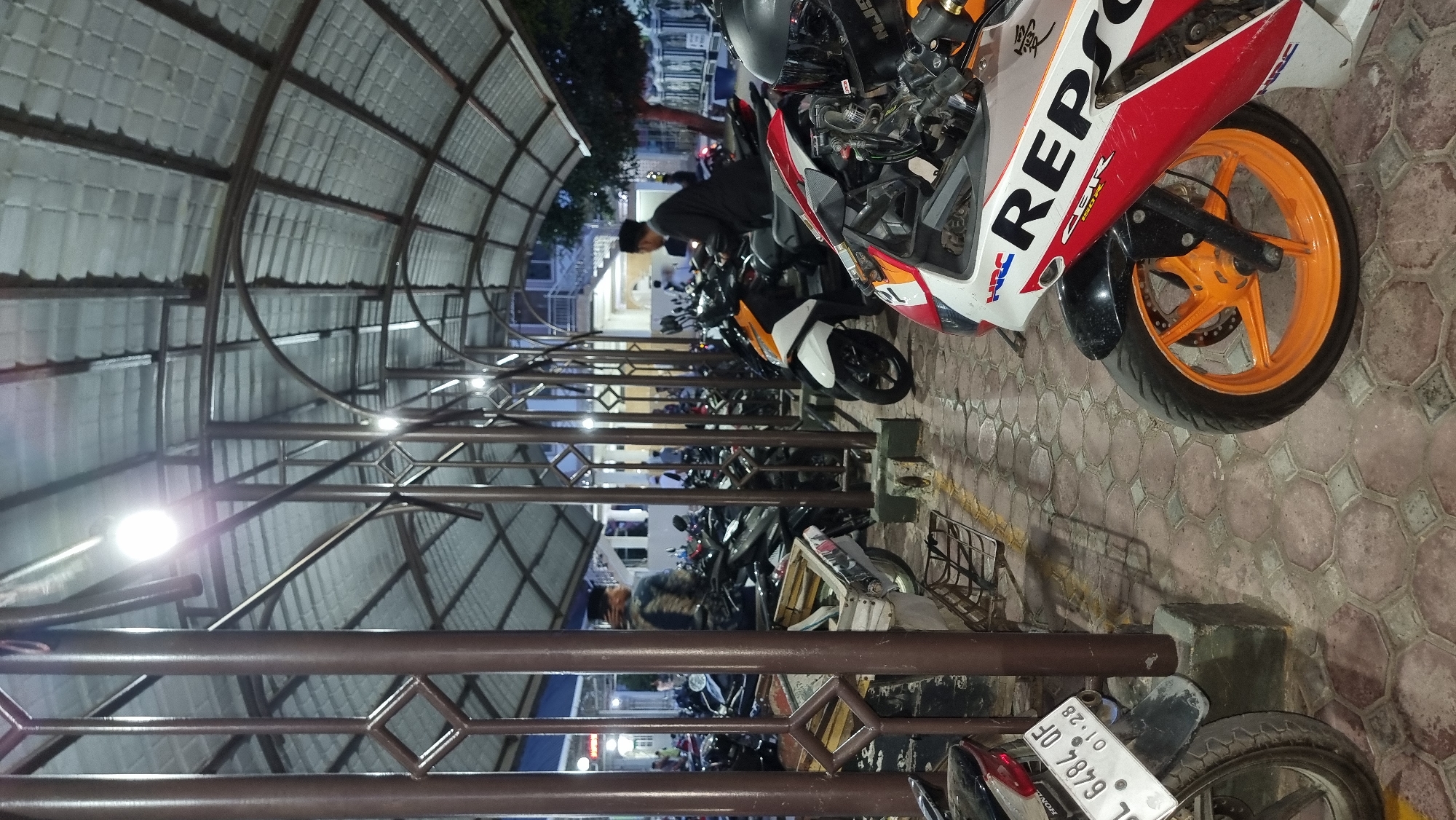 The image depicts a motorcycle parking area in Keudekruenggeukueh, Indonesia, at dusk or night. The parking area is covered by a corrugated metal roof supported by metal poles. Several motorcycles are parked in rows. In the foreground, a white, red, and blue sport motorcycle with "REPSOL" branding is visible, featuring an orange rim. To its left, another motorcycle's rear is partially visible, with a license plate reading "BL 6484 QF 01-28". In the middle ground, a man wearing a dark jacket and a peci (a brimless cap) is seated on an orange and white motorcycle. Further back, another individual in dark clothing and a peci stands near a motorcycle, and other people are faintly visible in the background, suggesting activity within or near a building. The ground is paved with hexagonal tiles. The lighting appears to be artificial, with overhead lights illuminating the scene.