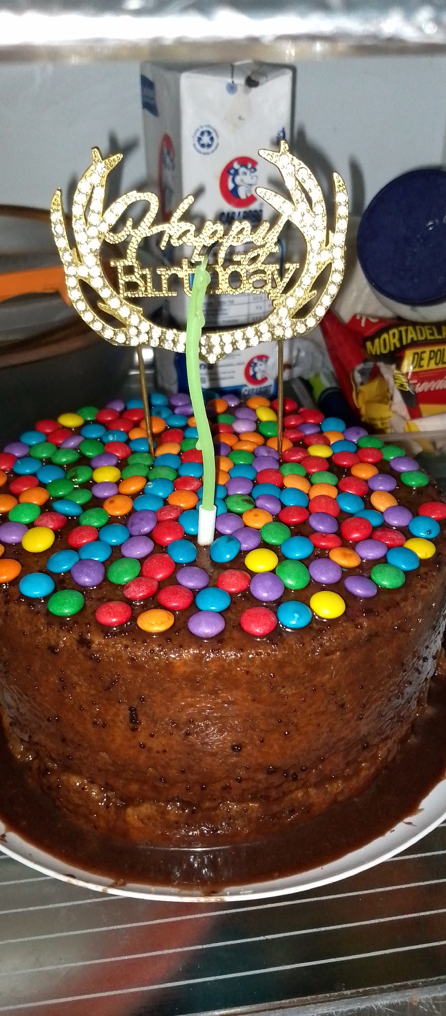 The image shows a close-up, eye-level shot of a chocolate cake decorated with colorful candies. A gold "Happy Birthday" cake topper adorned with rhinestones is prominently placed on top, with a single, unlit green candle emerging from the center of the cake. The cake itself appears to be moist and covered in a rich chocolate frosting, with some of the candies slightly embedded into the frosting. In the background, blurred elements suggest an indoor setting, possibly a kitchen or dining area, with a white cardboard box and a red bag of what appears to be deli meat visible. The overall lighting is dim, with a slight yellowish cast. There are no people visible in the image. The image does not provide any specific information about its location in the city of Caracas, Venezuela.