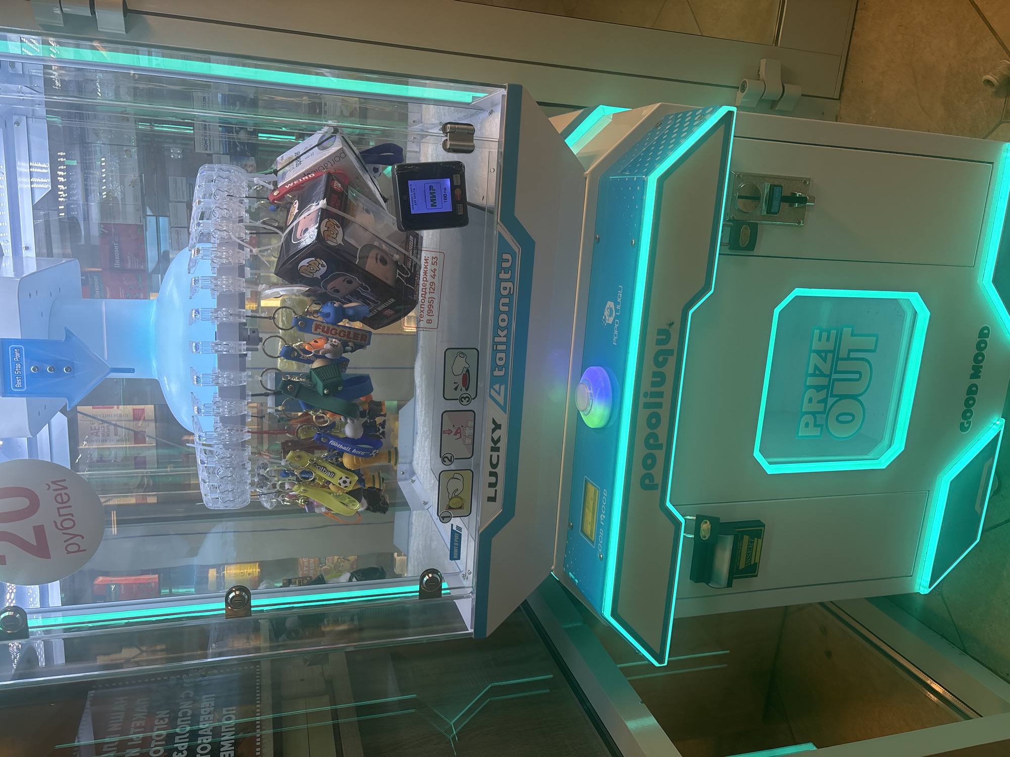 This is a medium shot of a claw machine arcade game, likely located in Novosibirsk, Russia, as indicated by the Cyrillic text on the prize labels and the payment terminal. The machine is white with bright teal LED lighting around its base and top. The front of the machine has a touchscreen display showing "GOOD MOOD" and a large, glowing green circular button. Above this, the brand name "popoliuqu" is displayed in white text.
Inside the glass enclosure, a claw mechanism hangs suspended above a selection of prizes. These prizes include numerous keychains with various designs like footballs, cartoon characters, and small plush toys. There are also several boxed collectible figures, some of which appear to be "Funko Pop!" figures. A small payment terminal is visible to the right, displaying a "MIR" logo and "100 RUB" (Russian Rubles), suggesting this is a payment option for the game.
The background of the machine is filled with products displayed on shelves, suggesting it's situated within a retail environment such as a convenience store or shopping mall. Some of these products have Cyrillic labels, further supporting the Russian location. A prominent price tag within the machine reads "20 рублей" (20 rubles). The overall environment is well-lit, with the bright LED lights of the arcade game being the dominant light source. There are no people visible in the image.