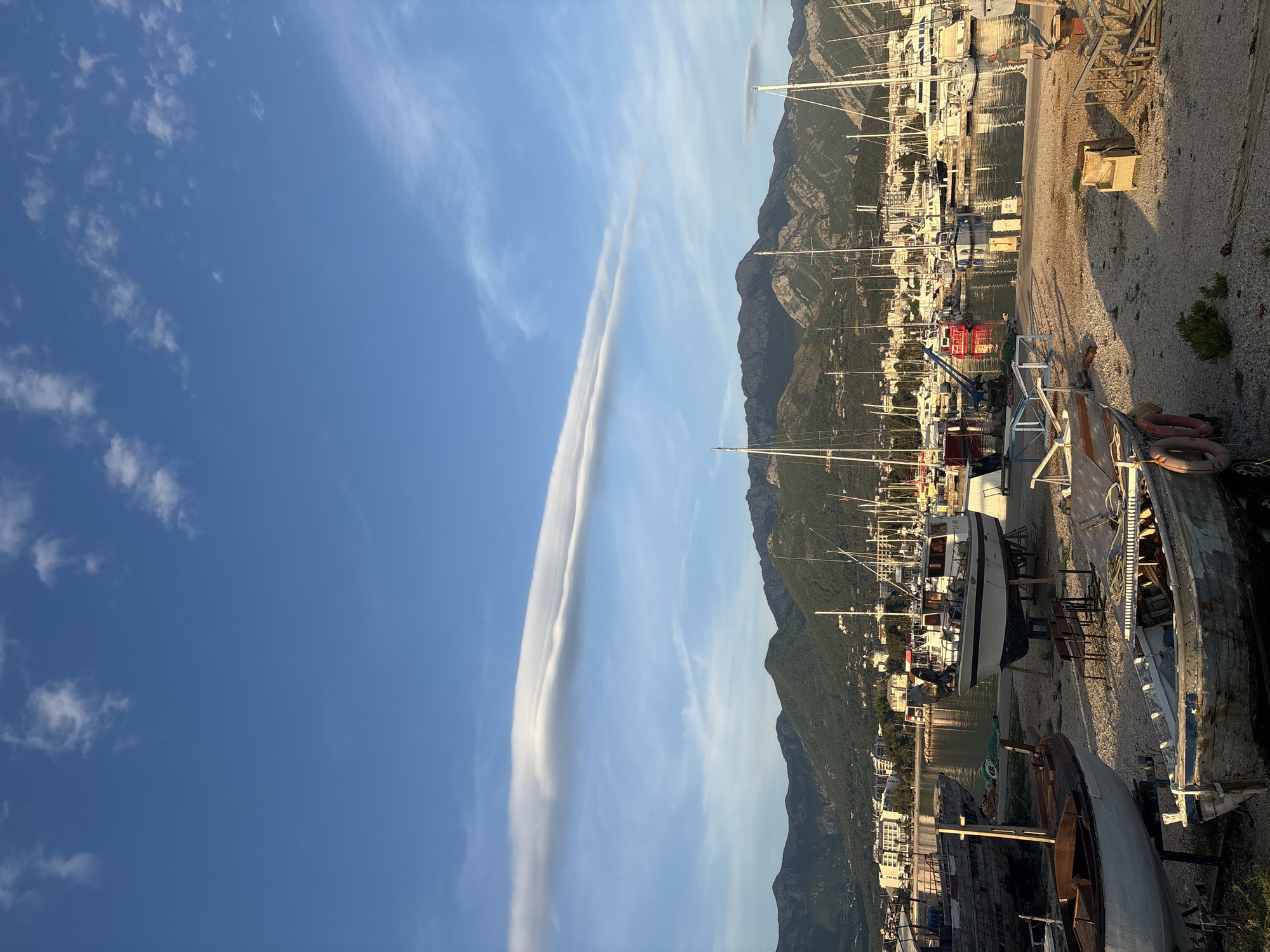 The image captures a vibrant harbor scene in Bar, Montenegro, under a clear blue sky. In the upper half, a long, distinct cloud formation, resembling a contrail or a lenticular cloud, stretches horizontally across the sky, creating a striking visual element. Scattered white cumulus clouds are also visible.
The background is dominated by a majestic range of mountains, covered in lush green foliage, extending along the horizon. Their rugged peaks contrast with the calm sky.
In the mid-ground and foreground, a bustling marina and shipyard are depicted. Numerous boats and yachts of varying sizes are docked in the water, their masts creating a dense pattern against the mountainous backdrop. The water reflects the boats and the sky. Along the gravelly shore, several boats are pulled out of the water, resting on supports, suggesting ongoing maintenance or storage. These include what appear to be older, traditional wooden fishing boats alongside more modern vessels.
The foreground also shows various elements typical of a working harbor: orange life preservers, ropes, metal structures, and what looks like a wicker chair. The lighting indicates either early morning or late afternoon, with long shadows and warm sunlight illuminating parts of the scene. No discernible text is visible.