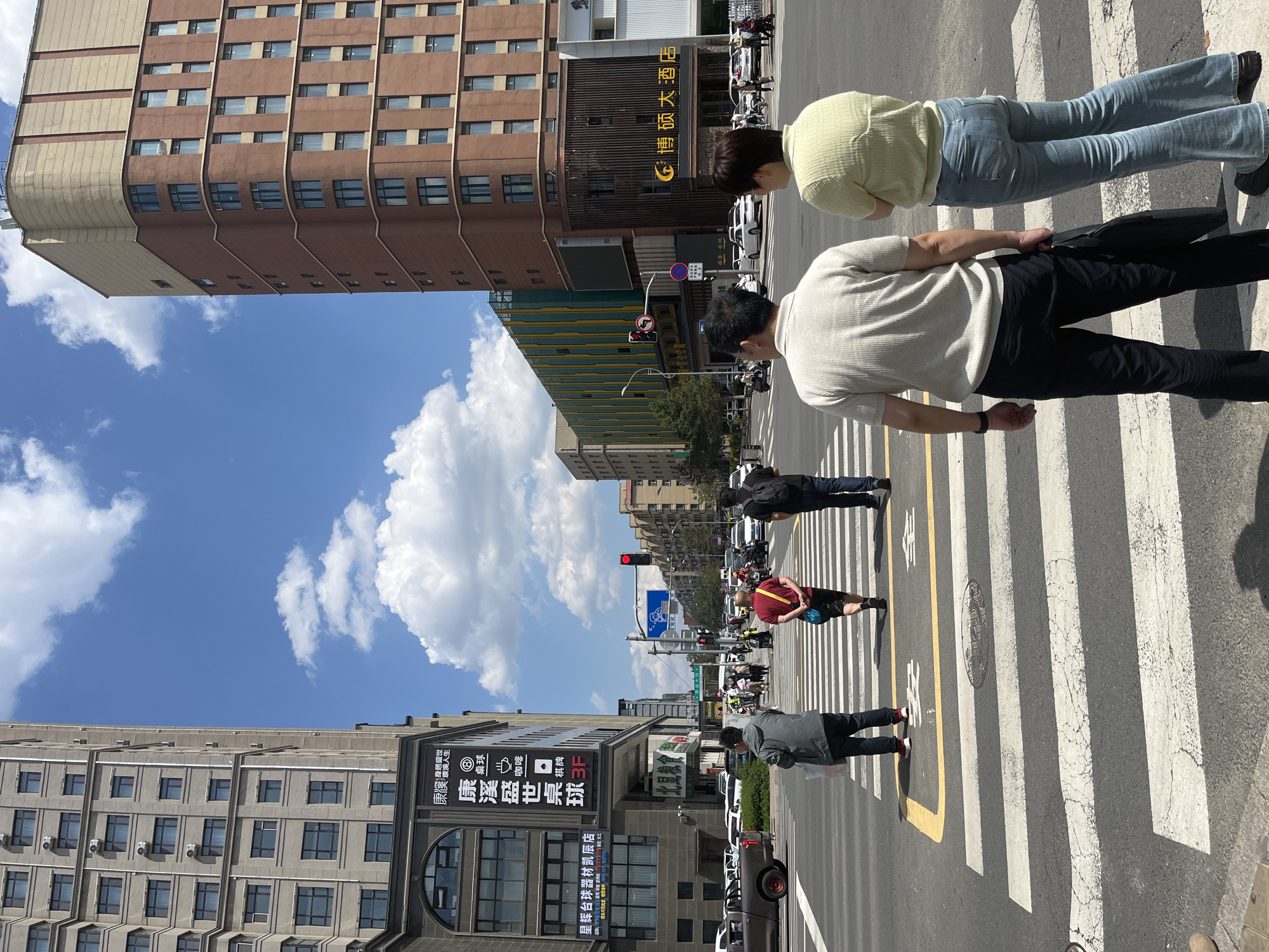The image shows a city street in Changchun City, China, featuring pedestrians crossing a crosswalk. Two individuals, a man and a woman, are closest to the camera and are in the foreground. Several more people are seen further down the street, also crossing. Buildings flank both sides of the street, including one building with an advertisement for "Kangxi Life" and a sign for the "Bosuo Grand Hotel". Cars and scooters are parked along the street. The sky is blue with white clouds. A traffic light is visible in the distance, displaying a red signal.
