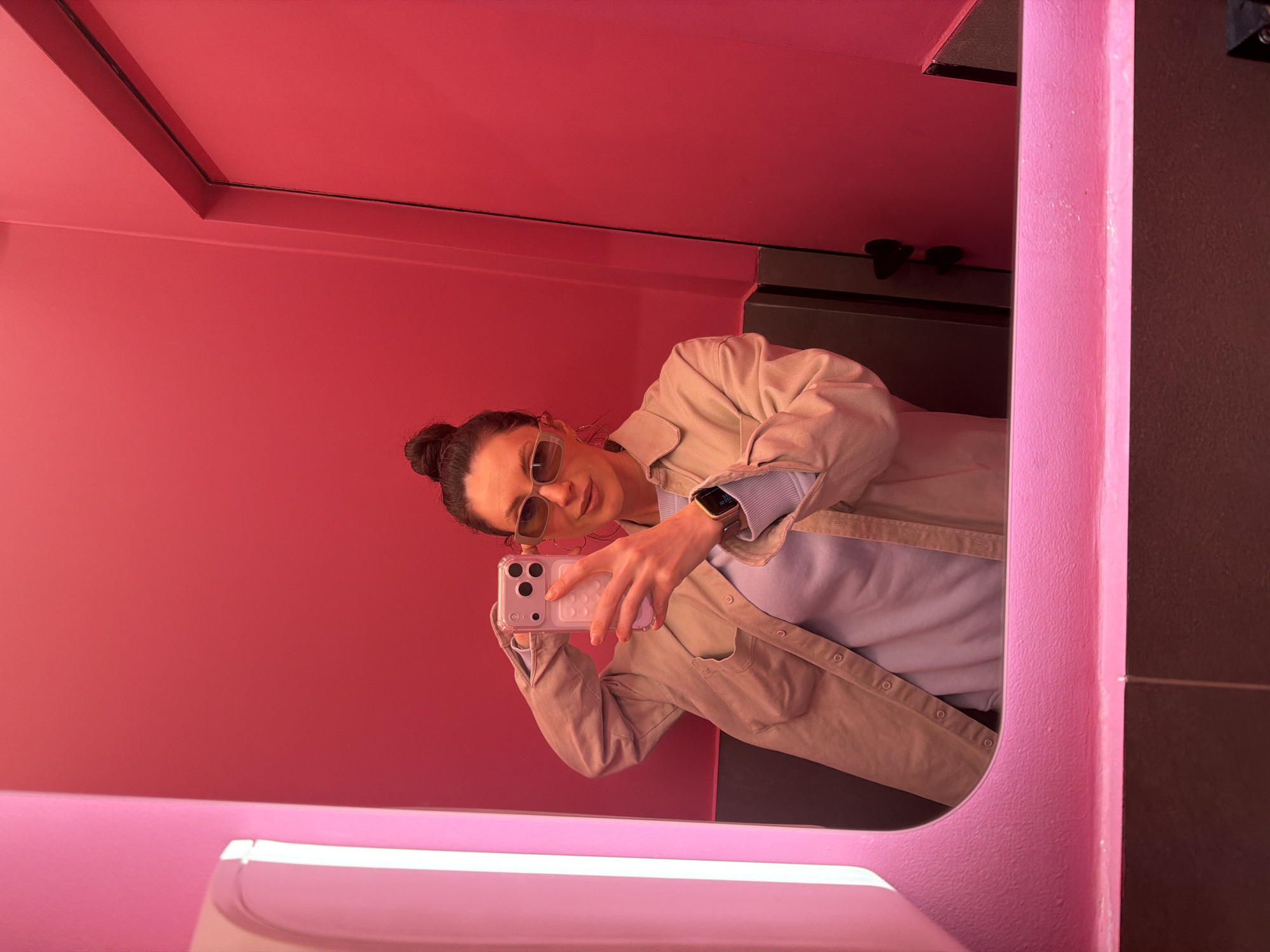 A woman is taking a selfie in the mirror of a pink bathroom. The location is likely in Perm, Russia. The woman has her hair in a bun and is wearing sunglasses, a light jacket, and a watch. She is holding a smartphone in her right hand. The bathroom is a uniform pink color and contains a sink and mirror.