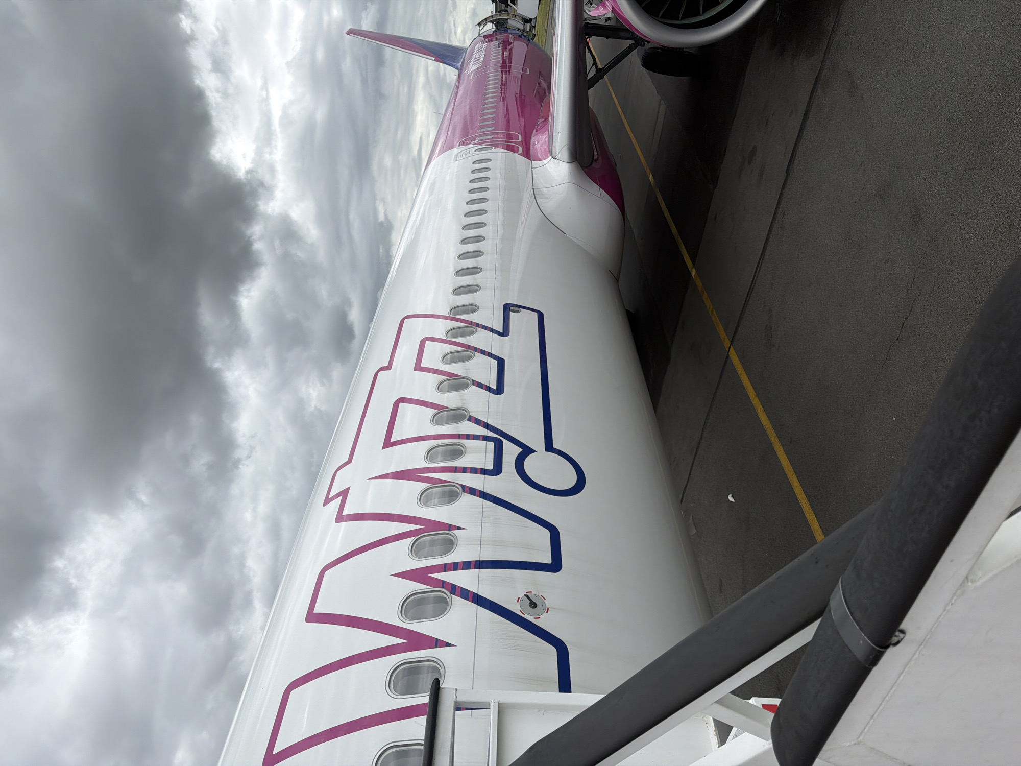 A Wizz Air airplane is depicted parked on an airport tarmac under a heavily overcast, grey sky. The image is taken from a low-angle perspective, looking up and along the side of the aircraft's fuselage. The plane's livery is primarily white, adorned with a large, stylized "Wizz" logo outlined in magenta and blue on the fuselage. The rear section of the fuselage and the vertical stabilizer (tail fin) are solid magenta, with "Wizz Air" text visible on the tail.
Numerous passenger windows are visible along the length of the fuselage. In the upper right of the frame, a portion of one of the aircraft's engine nacelles can be seen. The ground beneath the plane is dark grey tarmac, marked with yellow lines. A structure resembling boarding stairs or a jet bridge is present in the foreground, connecting to the aircraft, suggesting either passenger boarding or disembarkation is underway or imminent. A small circular gauge or port is visible on the fuselage near the blue part of the "Wizz" logo.
The weather appears to be a cloudy day, with no direct sunlight. No people are explicitly visible. The scene captures an industrial aviation environment, likely at an airport in Tillé, France.