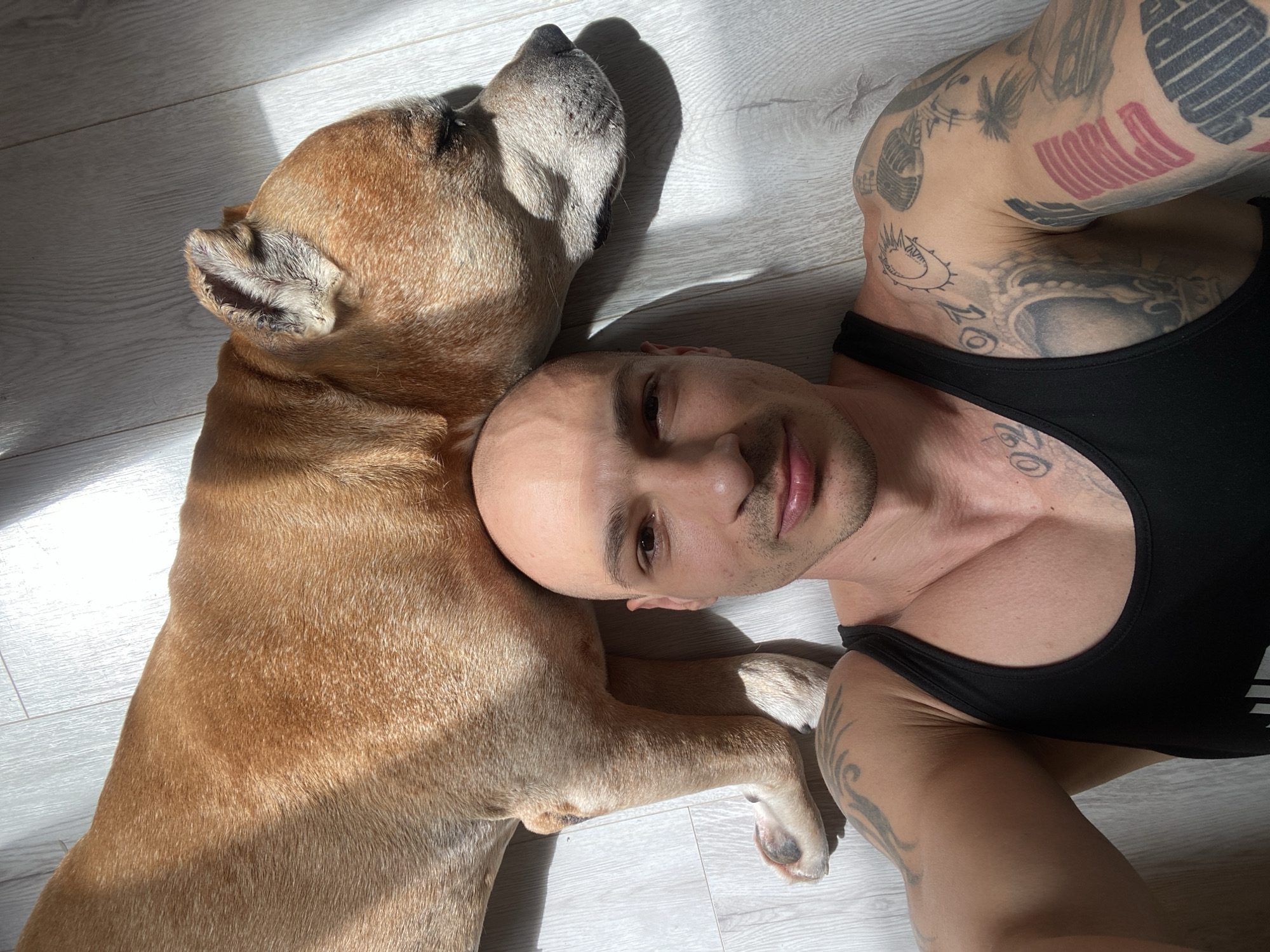 A light-skinned, bald man in a black tank top lies on a light gray wood floor next to a tan dog. The dog rests its head on the man's head. Both are in the sunlight. The man has many tattoos on his arms. The location is unspecified, but likely indoors and possibly in Pleven, Bulgaria.