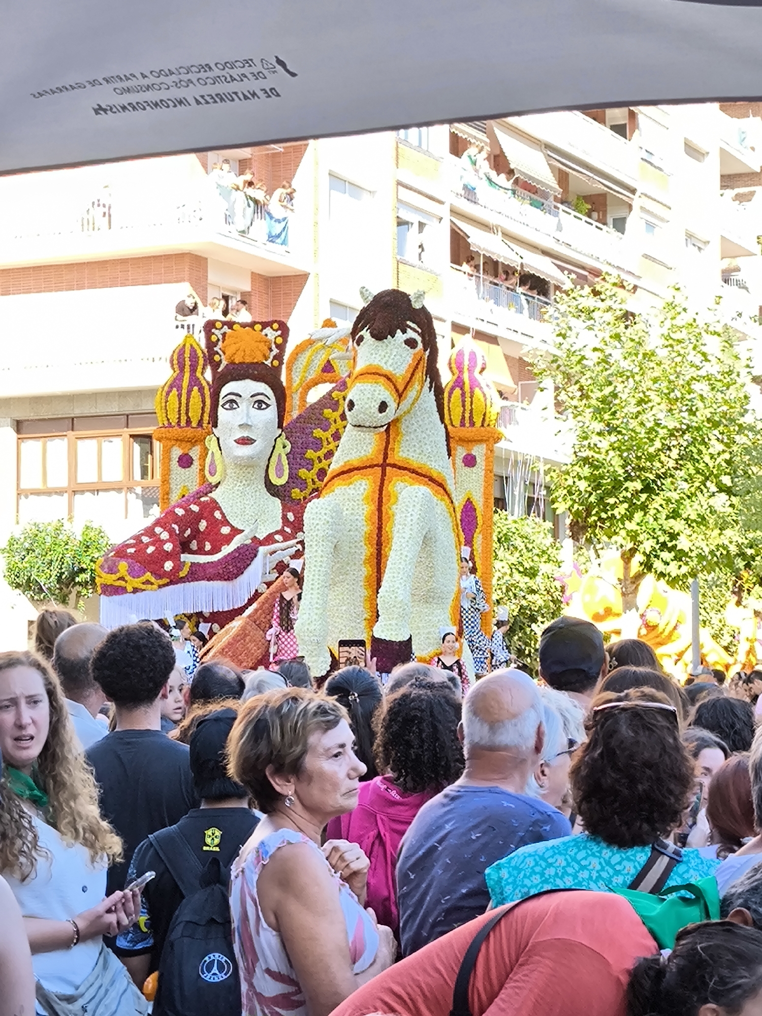 The image depicts a festive outdoor event, likely a parade, in Laredo, Spain. The central focus is a large, elaborate float adorned with flowers, featuring a prominent sculpture of a woman and a horse. The woman's face is white with painted features, and she wears a red and white polka-dot garment with yellow and purple floral accents. Beside her, the white horse, also decorated with yellow and orange flowers, appears to be part of the same float structure.
A crowd of people, dressed in casual summer clothing, are gathered around the float, observing the spectacle. Many are looking towards the float, with some holding up phones to take pictures or videos. The attendees' attire suggests a warm, sunny day.
In the background, buildings with balconies and awnings are visible, indicating an urban setting. Green trees and lush foliage are interspersed, adding to the lively atmosphere. The overall impression is one of a vibrant community celebration.