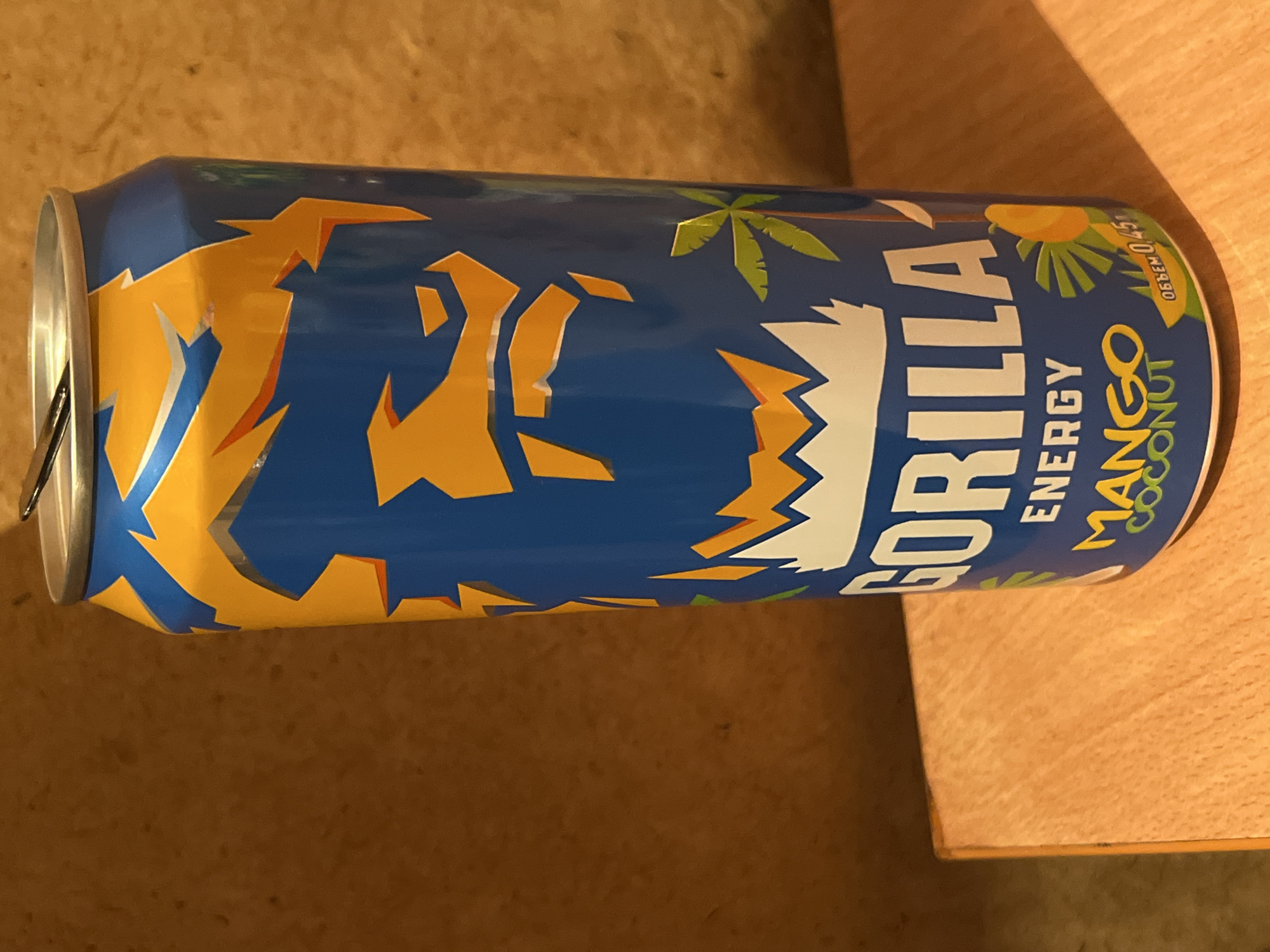 This is a close-up, eye-level, medium shot of a blue and yellow energy drink can with the brand name "GORILLA" prominently displayed in large white letters. The can also features images of a gorilla's face rendered in yellow shapes against the blue background, as well as palm trees and a mango. Below "GORILLA," "ENERGY" is written in smaller white letters, followed by "MANGO COCONUT" in yellow letters with a green outline. The bottom right of the can shows the text "ОБЪЕМ 0,45". The can is positioned on a light brown wooden surface, likely a table. The background is a textured brown surface. There is no indication of the location being in Iksha, Russia.