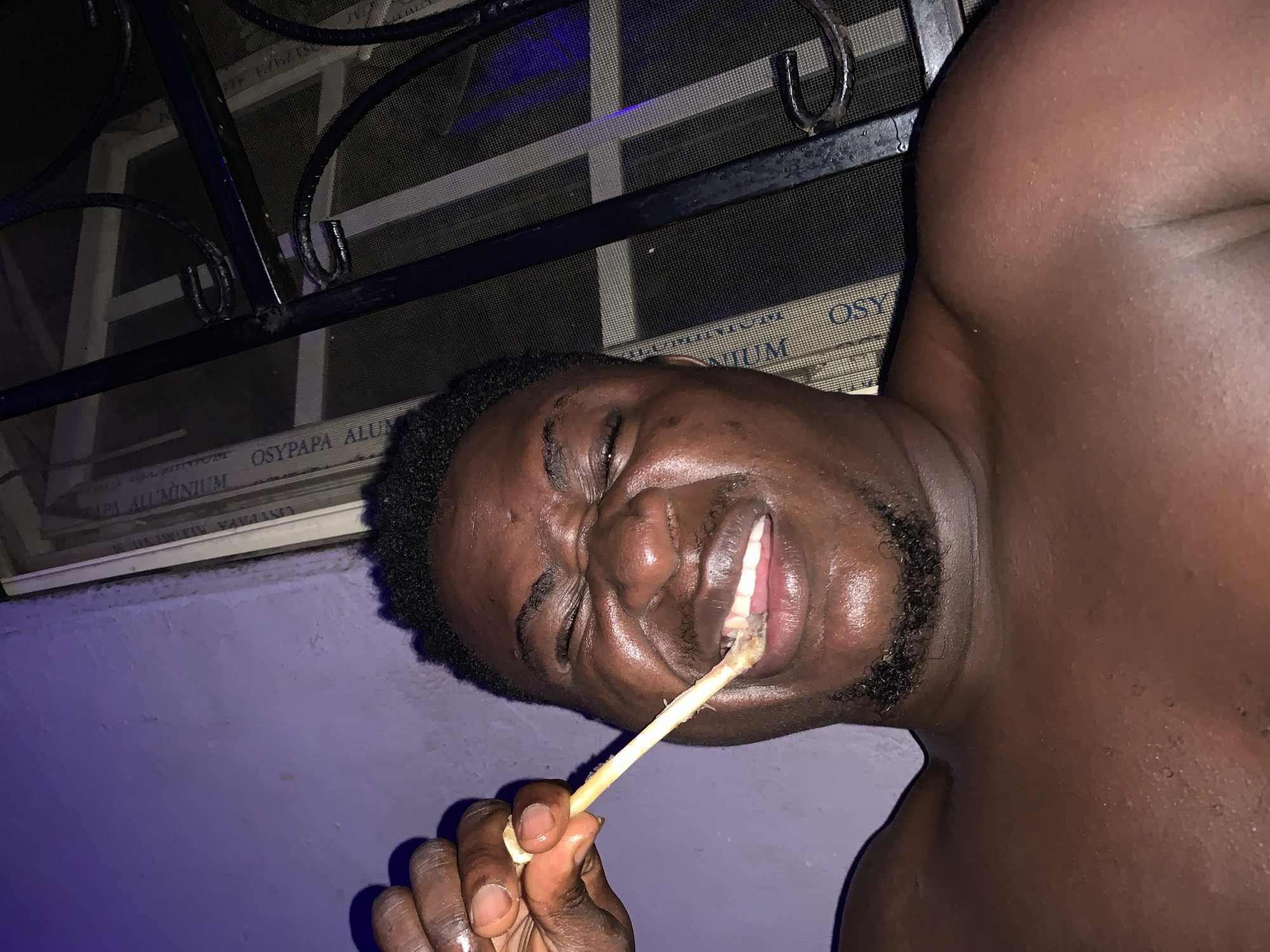 An image, apparently taken in Akwanga, Nigeria, features a shirtless Black man lying on his side, with a screen door with black wrought iron in the background. He is holding what appears to be a chicken bone in his hand, with a portion of it in his mouth. His eyes are closed and his face appears contorted in what could be interpreted as a grimace. His skin glistens, likely with sweat or oil. The lighting suggests an indoor environment illuminated by artificial light. The wall is visible in the background.