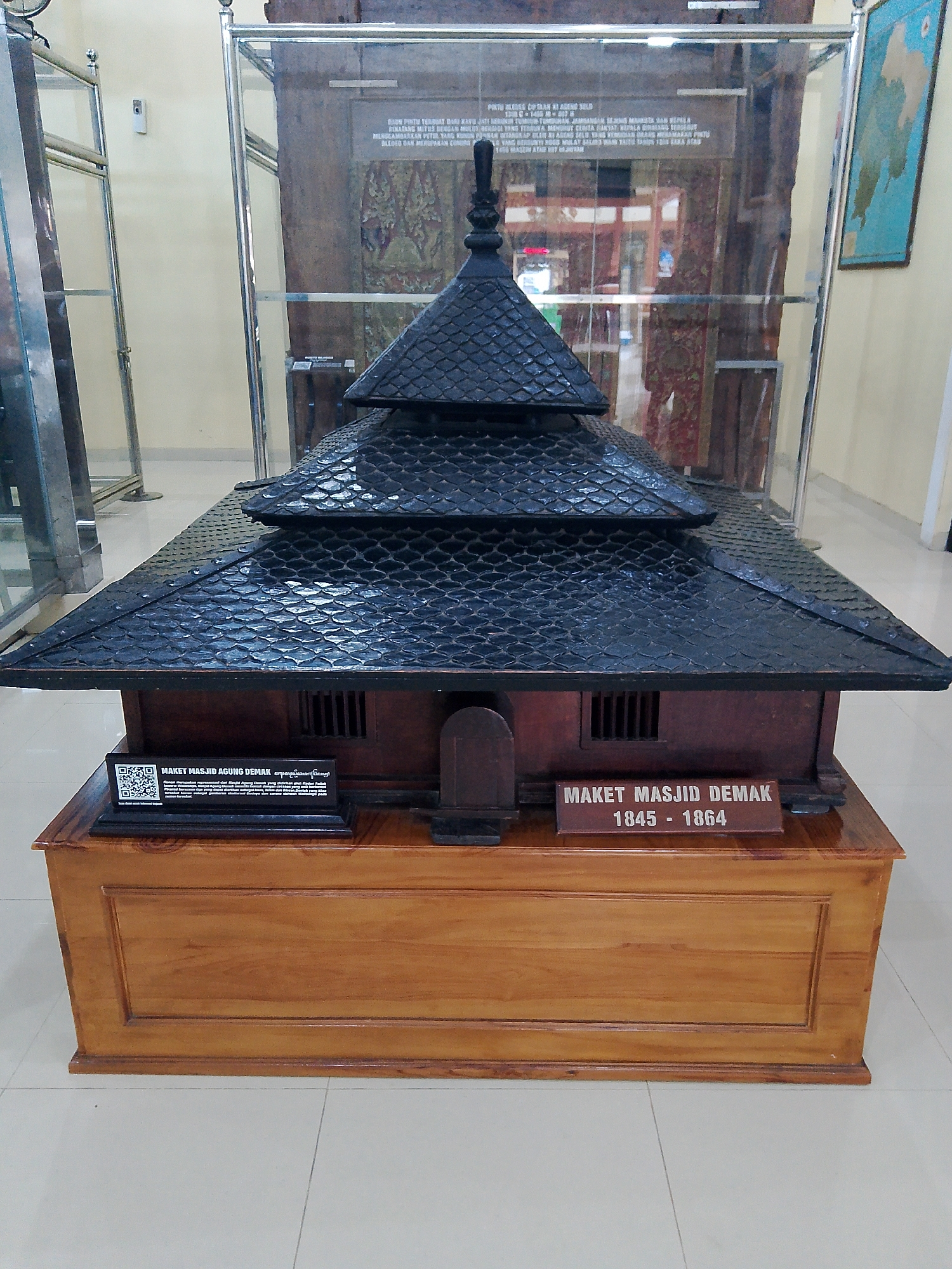 A detailed model of the Demak Grand Mosque is displayed in what appears to be a museum exhibit. The model, constructed primarily from dark wood with a black tiled roof, features multiple tiered roofs culminating in a finial. The base of the model is a polished wooden pedestal with a plaque indicating "MAKET MASJID DEMAK 1845 - 1864" (Model of Demak Grand Mosque 1845-1864). A QR code and additional text are visible on a sign in front of the model. The exhibit is enclosed in a glass display case, with other museum artifacts, including a framed map and possibly fabric hangings, visible in the background. The surrounding environment suggests an indoor public space, likely a museum, with tiled flooring and neutral-colored walls. There are no people visible in the frame. The overall lighting is even, highlighting the model and its display.