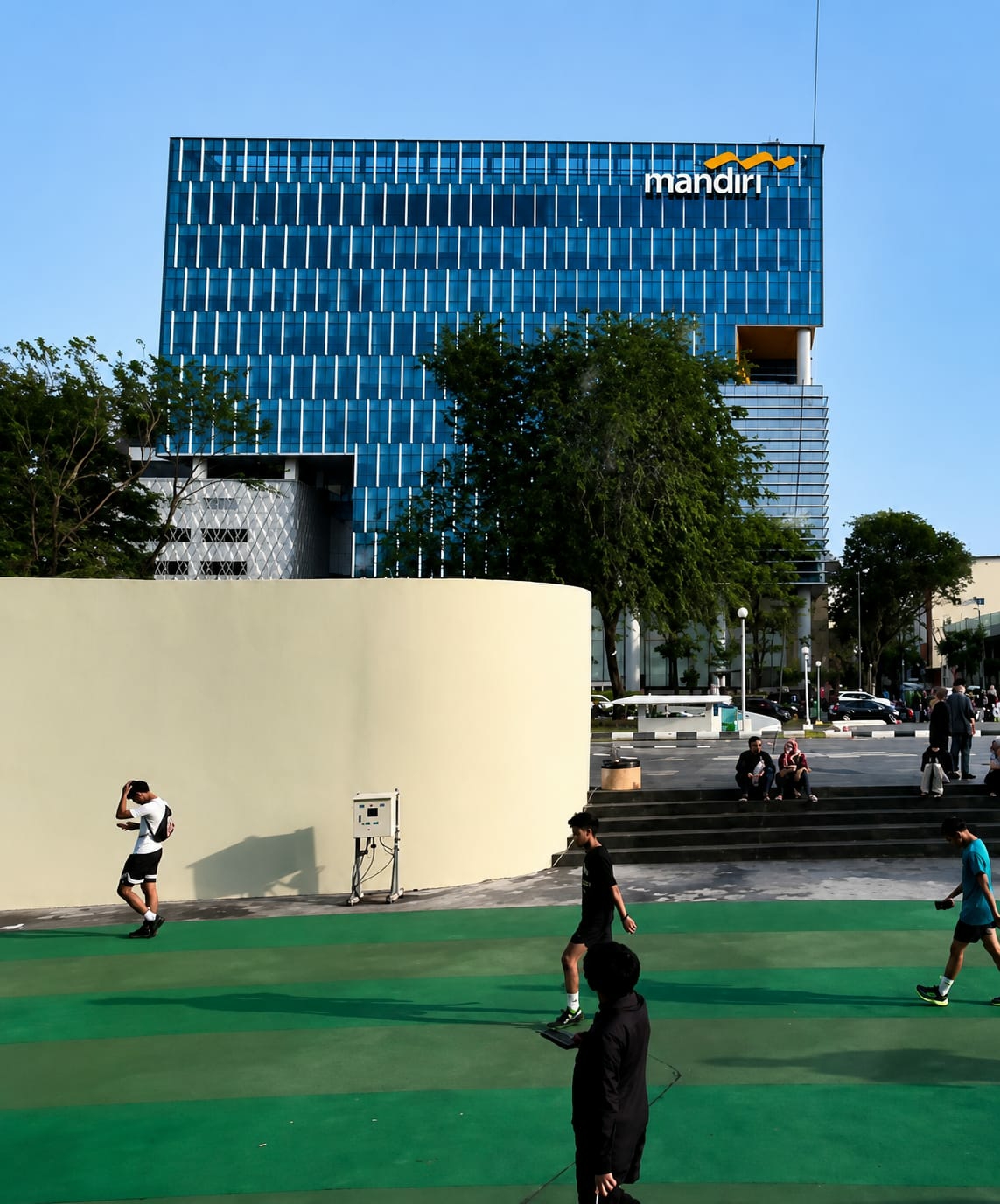 In the City of Medan, Indonesia, an outdoor urban scene unfolds beneath a clear blue sky. Dominating the background is a modern, multi-story building with a striking blue glass facade, distinguished by white vertical lines and the orange "mandiri" logo at the top. In the mid-ground, a light-colored, curved wall forms a prominent feature, casting a distinct shadow. In front of this wall, an individual in athletic wear walks with their hand on their head, while a short distance away, another person in dark attire jogs. Further back, a white electronic device on a stand is positioned near the wall. The foreground is characterized by a green surface, segmented by lighter green stripes, indicating a recreational or sports area. Several people are visible on steps to the right of the curved wall, some seated and others standing, with a few walking on the green area. The overall environment suggests a public space with a blend of architecture and open areas.