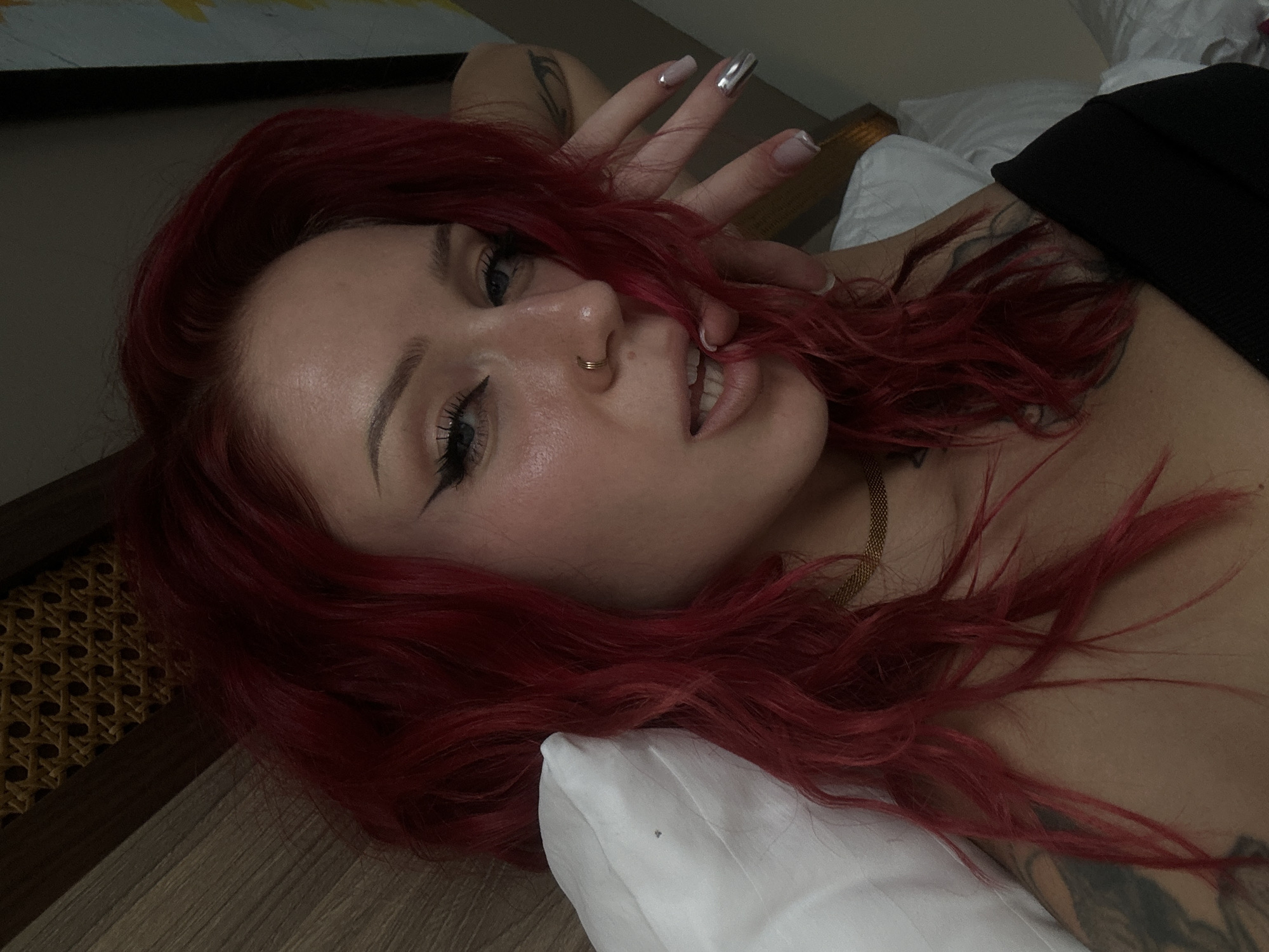 A woman with red hair lies on a white pillow, her head resting on a wooden surface. She has a nose ring and is wearing makeup, including winged eyeliner. Her hand is near her face, showcasing long, manicured nails. Part of her chest and neck are visible, adorned with a gold chain. The background includes a painting and white bedding. The location appears to be indoors, possibly a hotel room, but no information from the city of Unknown, Indonesia is present.