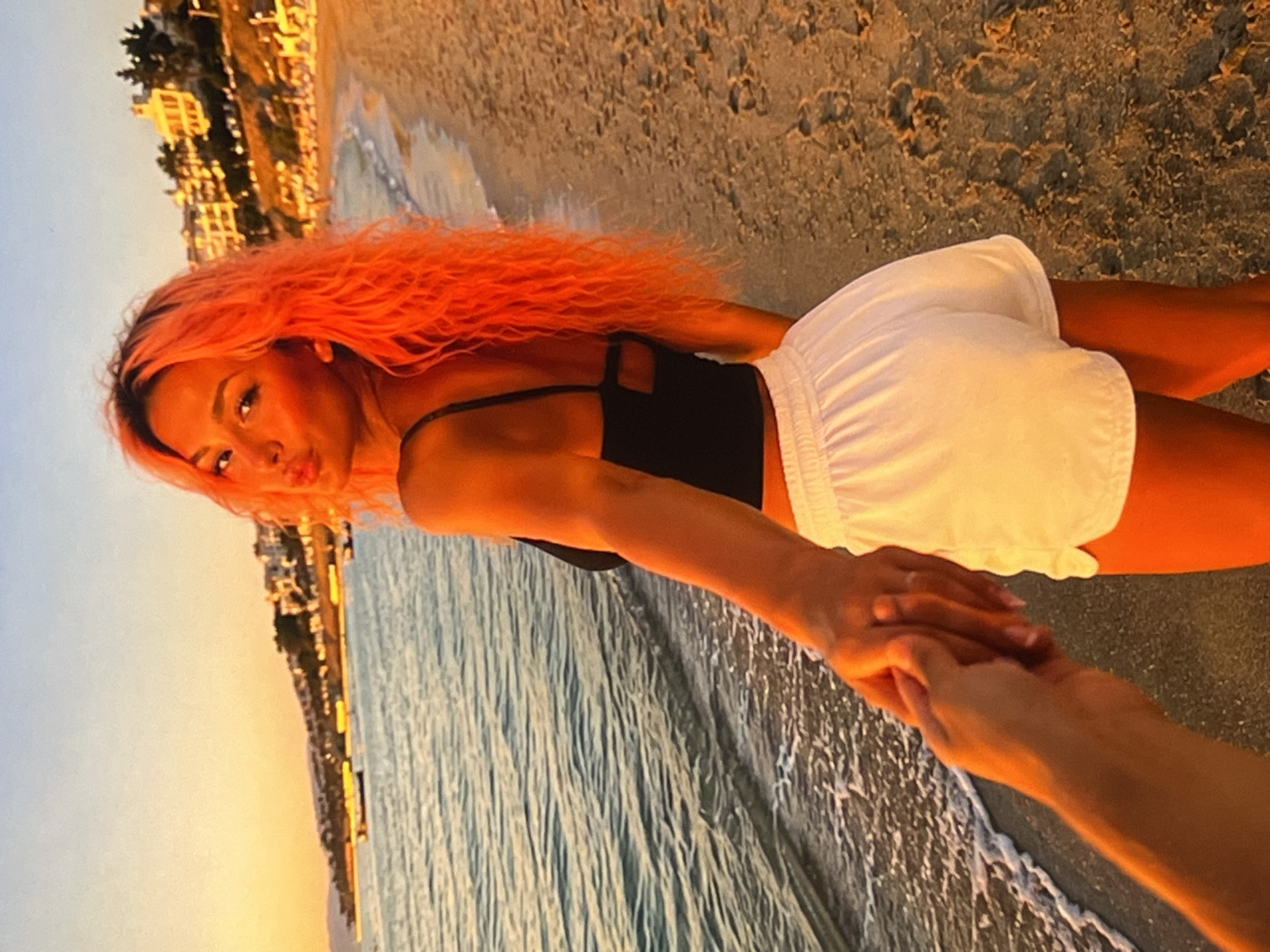A first-person perspective shot shows a person's hand holding another person's hand. The second person, facing away from the camera and looking over their left shoulder, is walking on a sandy beach at sunset. They have bright orange, wavy hair and are wearing a black crop top and white shorts. The ocean is on the left of the frame, with gentle waves lapping the shore, and buildings are visible on a hill in the background.