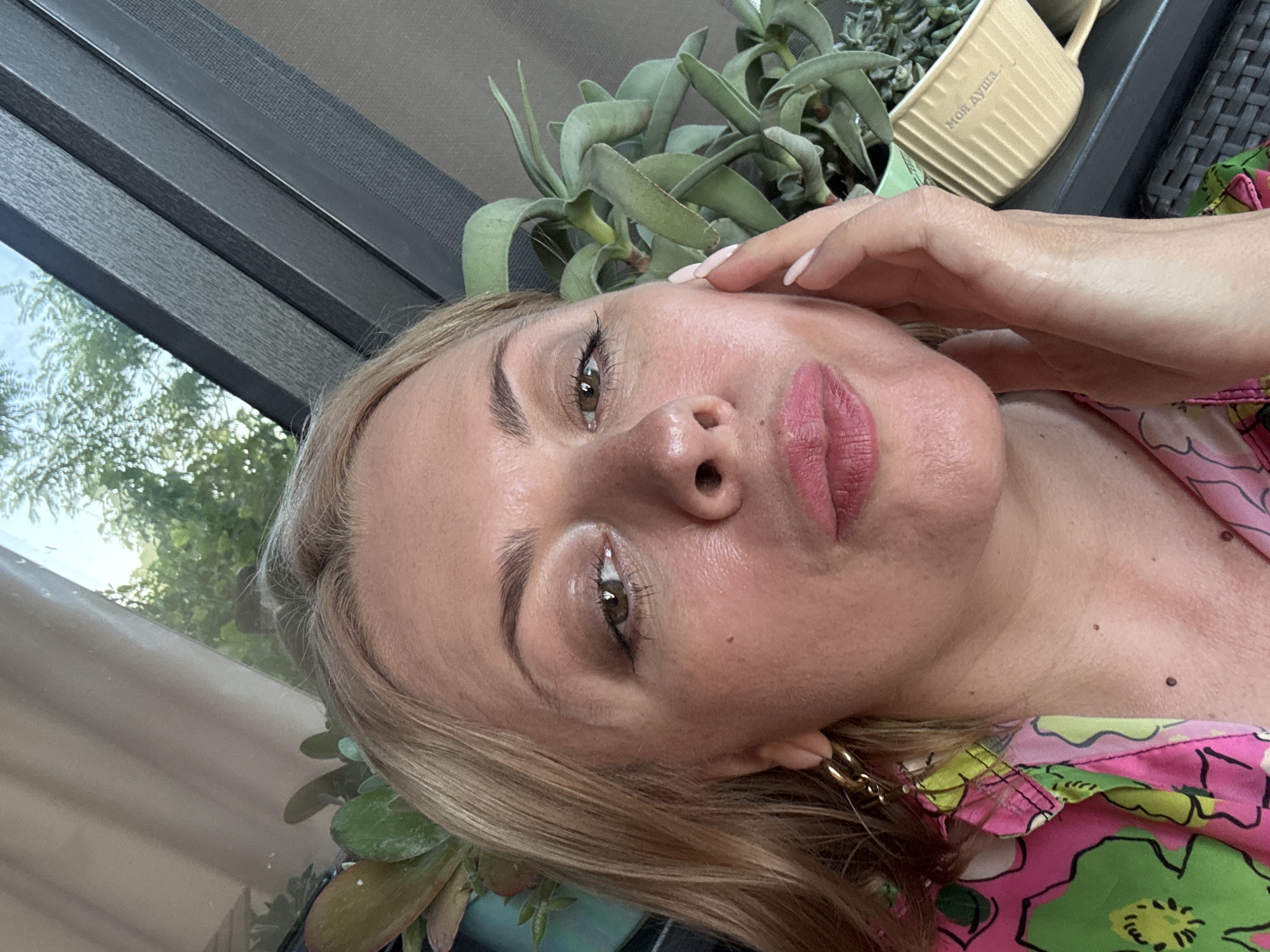 The media file is a close-up, low-angle selfie of a woman with blonde hair and fair skin. She is wearing pink lipstick and a colorful floral-print top in shades of pink, green, and yellow. Her right hand is touching her face near her eye. She is wearing a large gold hoop earring.
In the background, there is a window with a view of green trees and foliage, suggesting an outdoor setting. There are also some plants in pots, one of which is a succulent. A beige planter with embossed text in Russian, "Моя Душа" (My Soul), is visible behind her head. The lighting suggests it is daytime. The overall mood appears relaxed and casual. The location is identified as Sochi, Russia.