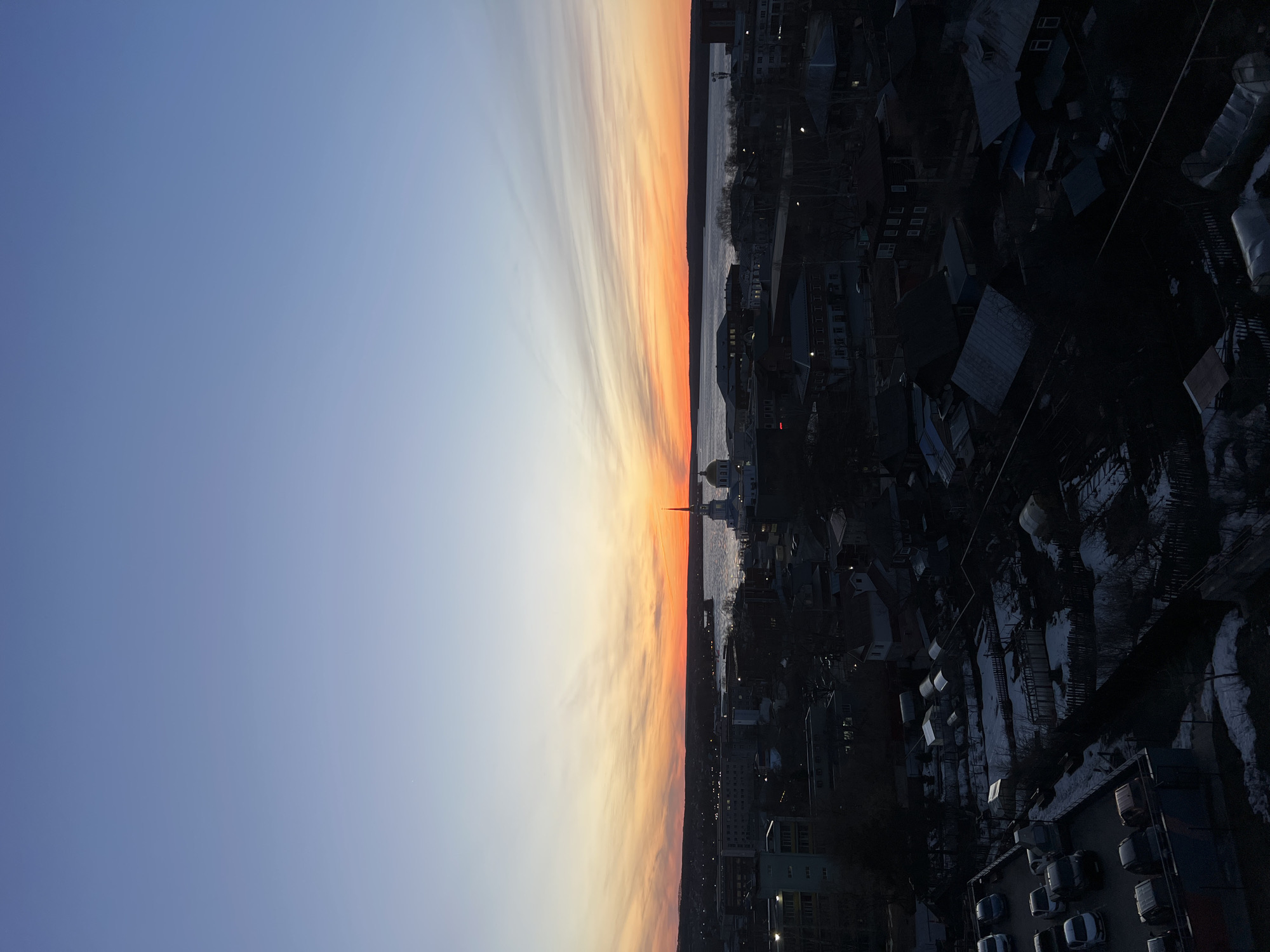 The image shows a cityscape during sunset. The sky transitions from blue to orange, with cloud formations stretching across the horizon. Below the sunset is a body of water that separates the sky from the city buildings. The cityscape is dominated by buildings of varying heights and styles, some with distinct rooftops. There's a cathedral with a golden dome and tall steeple visible. The overall impression is that of an urban environment transitioning from day to night.
Given the prompt's context, it is plausible to state the image is of Votkinsk, Russia. Specifically, the cathedral that is visible in the picture is most likely the Annunciation Cathedral, located in the center of Votkinsk. The picture is taken from above, as a drone, or from a high building looking at the city center and the Votkinsk water reservoir.