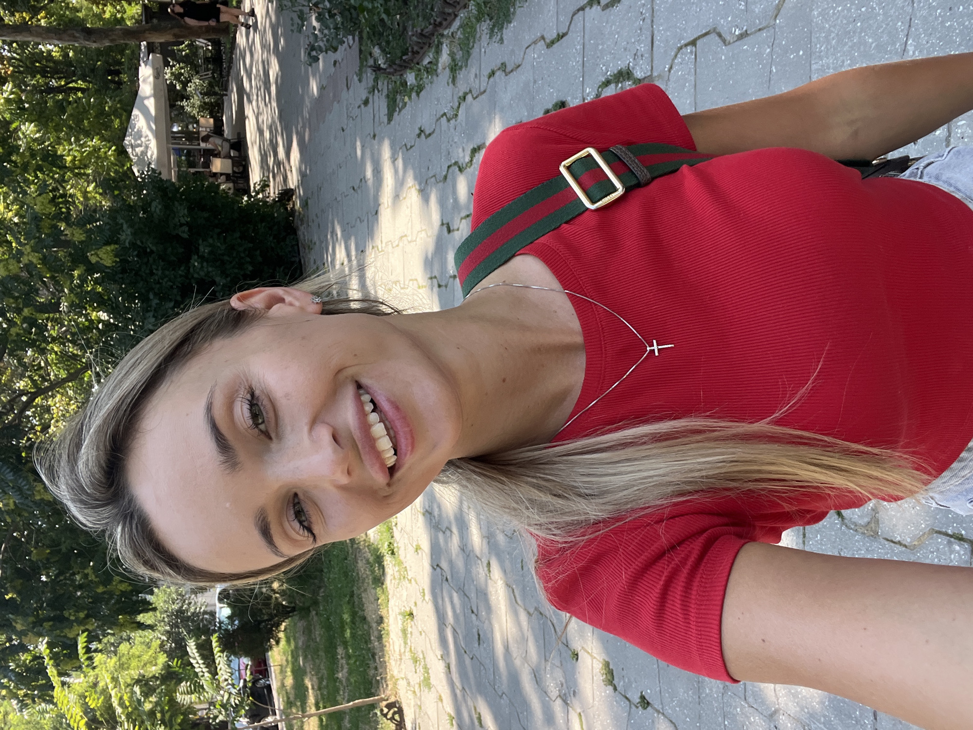 A blonde woman with fair skin, wearing a red t-shirt, light-colored shorts, and a silver necklace with a cross pendant, poses for a selfie outdoors. She has blonde hair pulled back in a ponytail and is smiling. She is carrying a bag with green and red stripes across her body. She is standing on a paved walkway with trees and greenery in the background. In the distance, there are people sitting at tables under an umbrella, suggesting a cafe or restaurant area. The sun is shining, casting shadows on the walkway. The location appears to be a park or a public area in the city of Odesa, Ukraine.
