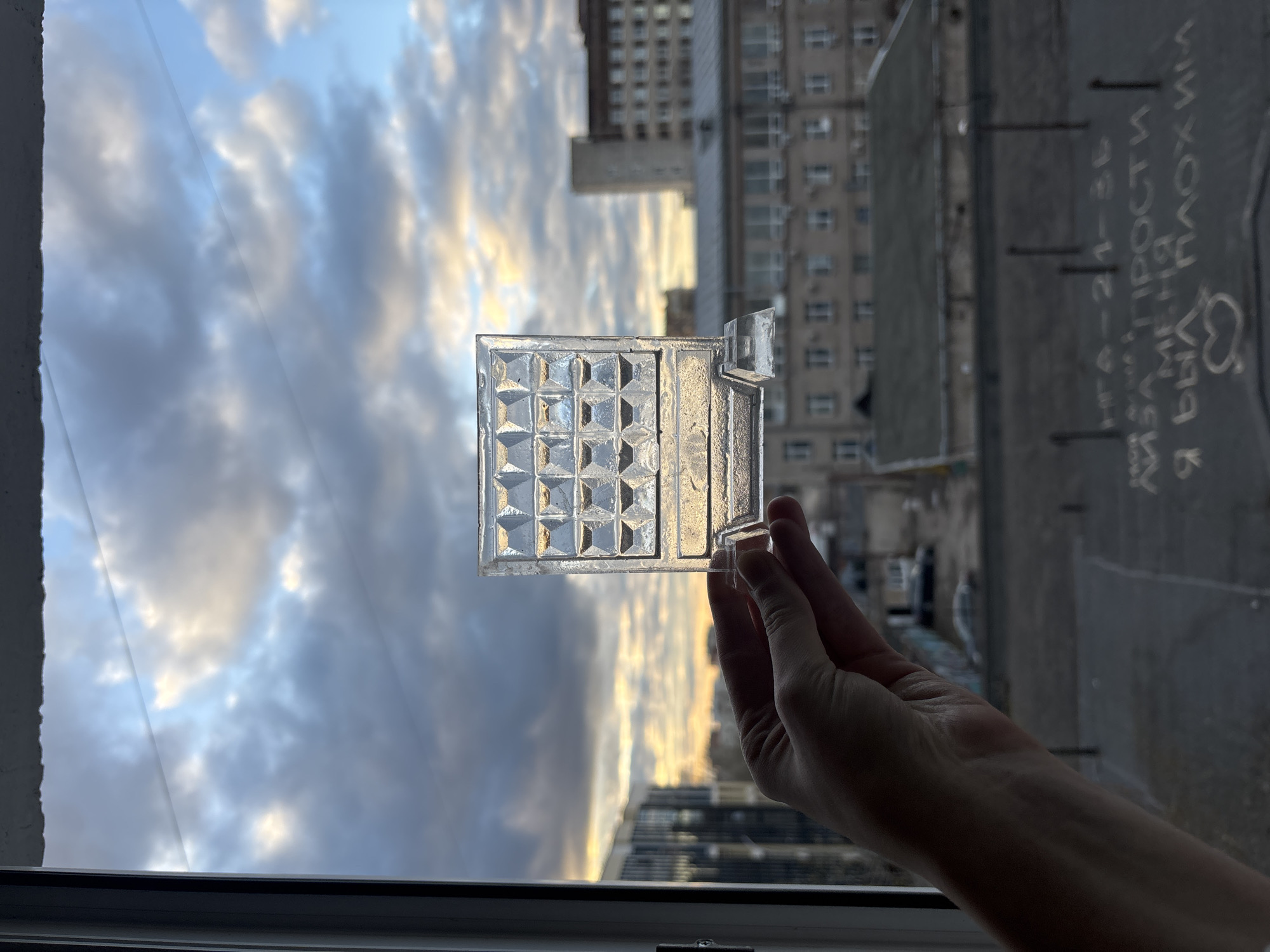 A hand holds a square, clear glass object with a grid pattern of raised squares in front of an open window. The background shows a cloudy sky and buildings in a city, likely Perm, Russia based on text on the wall: "Я бЫЛА ПЛОХАЯ" and "ПРА-21-36," along with other Cyrillic writing. The window frame is visible at the bottom of the picture.
