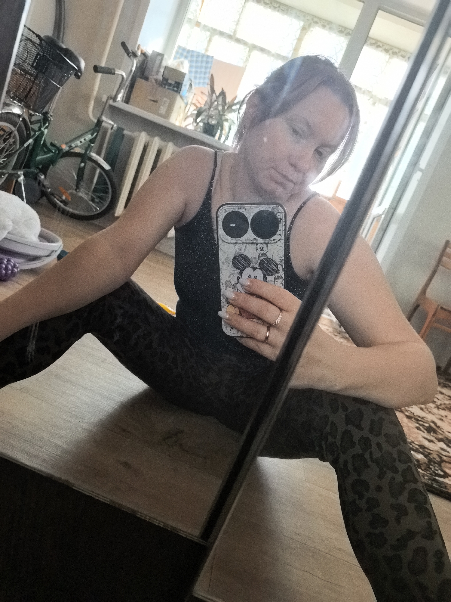 A woman is taking a selfie in a room that appears to be in Chaikovsky, Russia, possibly a residential setting or a studio. She is sitting on the floor, with her legs spread apart. She is wearing a sparkly black tank top and leopard-print leggings. In her right hand, she is holding a smartphone with a Mickey Mouse case. She is also wearing a ring on her ring finger.
In the background, there is a bicycle with a basket, a window with blinds, and some plants. The floor is wooden, and there is a rug on the right side of the image. The overall lighting suggests it is daytime. The room seems to be furnished with basic items, consistent with a functional living space.