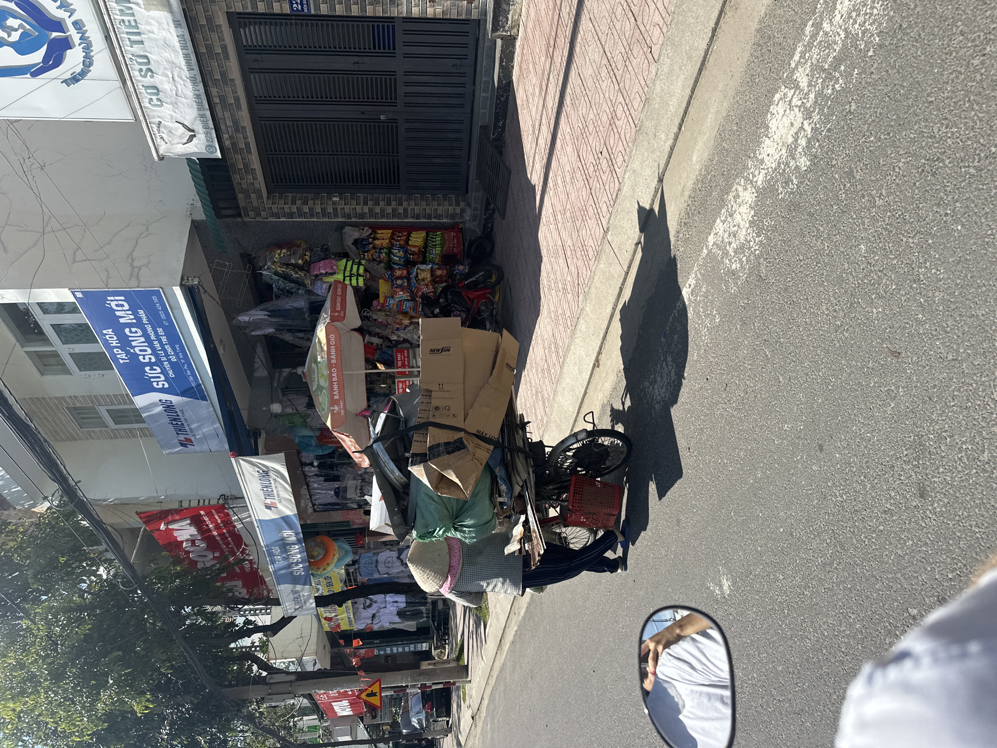 A person wearing a conical hat and plaid shirt, pushing a bicycle laden with a large quantity of cardboard boxes and green plastic bags, walks on the sidewalk alongside a street. The person is wearing dark pants and blue flip-flops. The bicycle has a red basket attached to the back. The scene takes place on a sunny day in front of shops, with awnings and signage visible. One sign reads "TẬP HÓA SỨC SỐNG MỚI" which translates to "New Life Grocery Store." Another sign says "CƠ SỞ TIỆM" and an address including "225 BIÊN PHÚ, VINH HOA, NT". This context indicates the location is likely in the city of Nha Trang, Vietnam. The street is paved and has a painted white line. In the foreground, a motorcycle side mirror reflects the arm of the person taking the photo. Trees and power lines are visible above the shops.