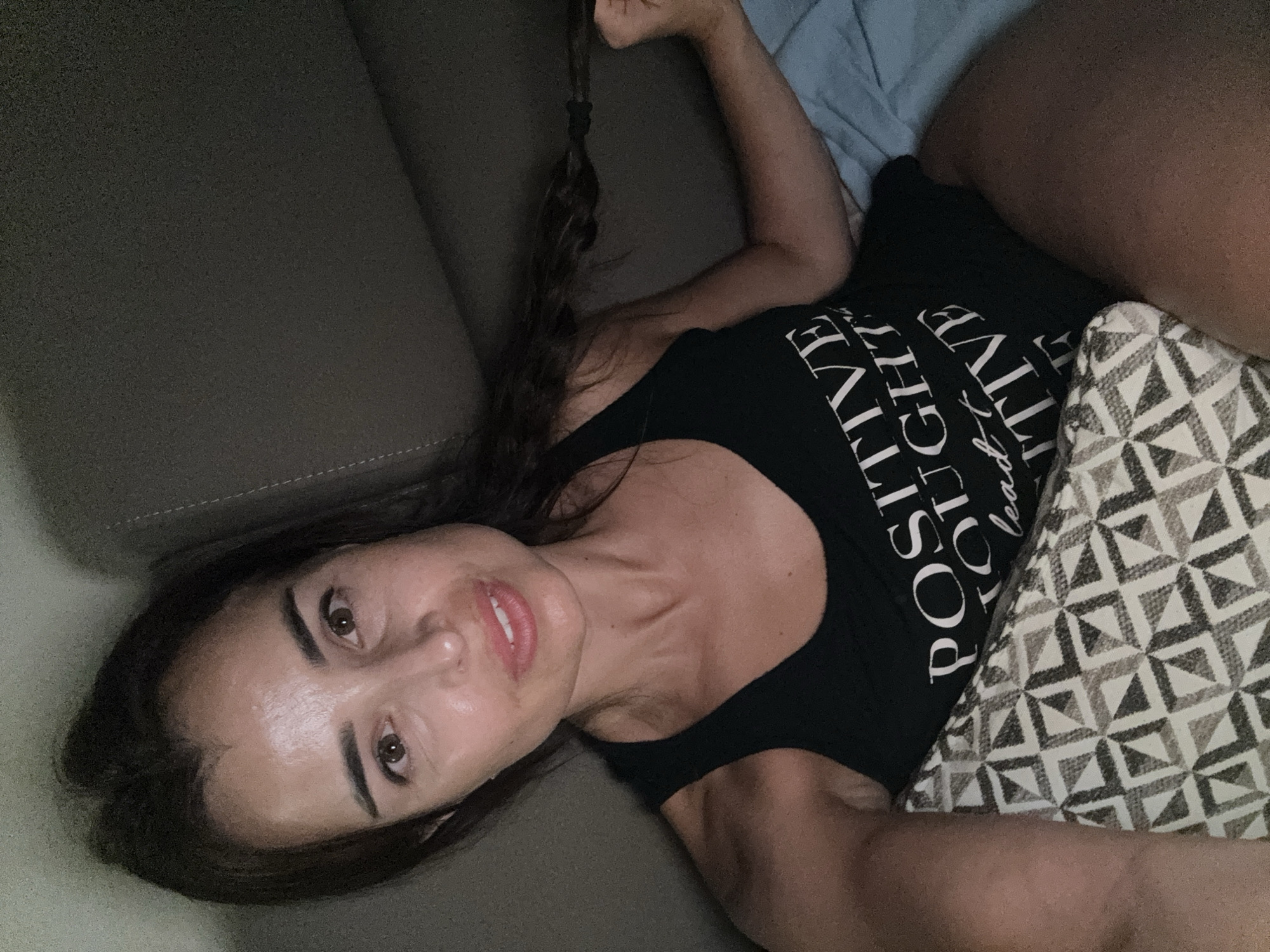 A woman with dark hair pulled back into a braid is lying on a couch, looking at the camera. She is wearing a black tank top with white text that reads "POSITIVE THOUGHTS LEAD TO POSITIVE". Her left arm is resting on a patterned pillow, and her right arm is visible holding the braid. The background is a dark couch and a blue blanket. The lighting is dim, suggesting an indoor setting at night.