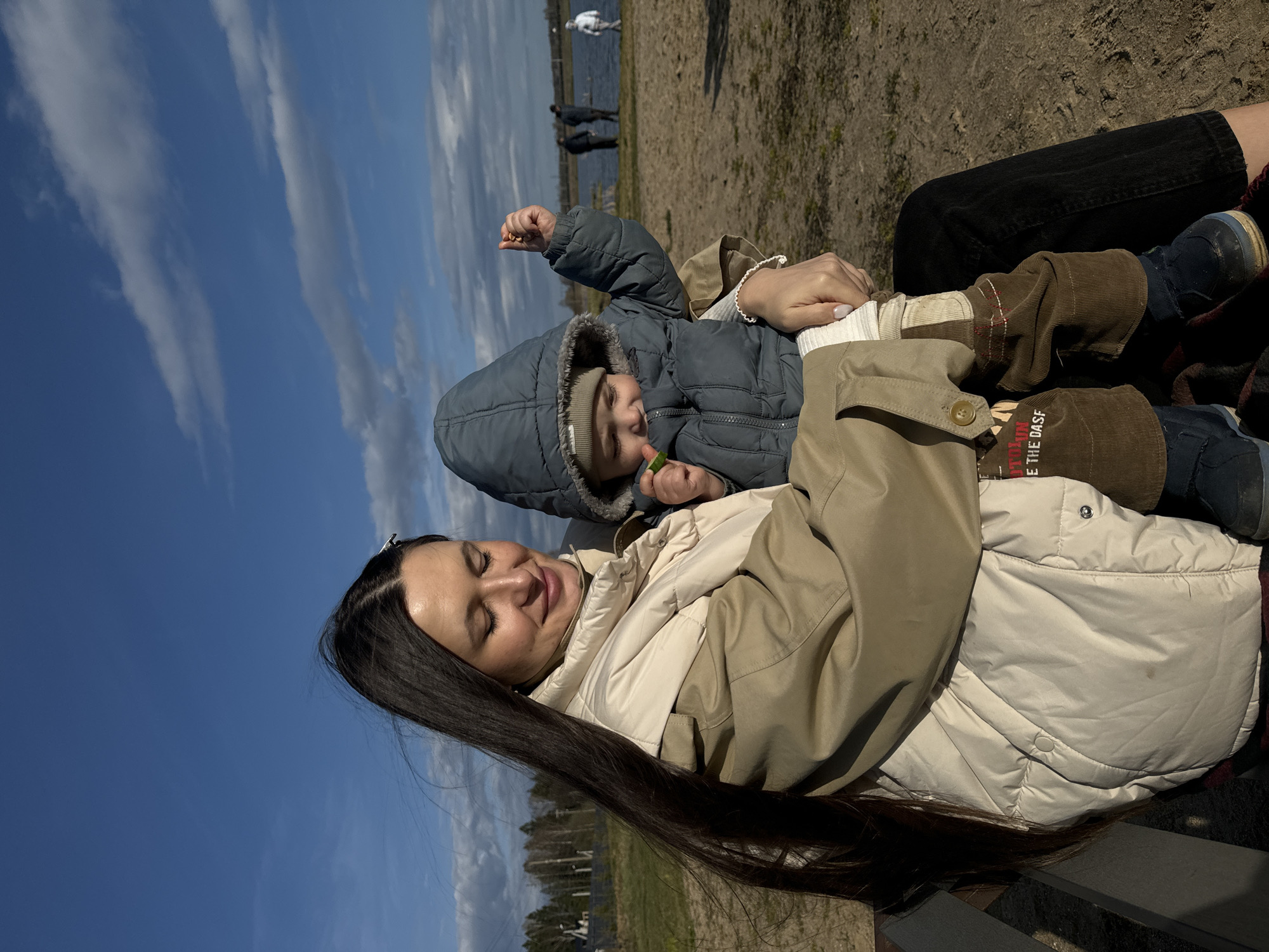 A woman and a toddler are sitting on a bench outdoors. The woman has long, dark hair and wears a light-colored puffer jacket. The toddler is wearing a blue hooded coat and brown pants. The toddler is holding food in their hand. In the background, there is a body of water, some people standing near the shoreline, and a cloudy blue sky. The image was taken in Литвиново, Russia.
