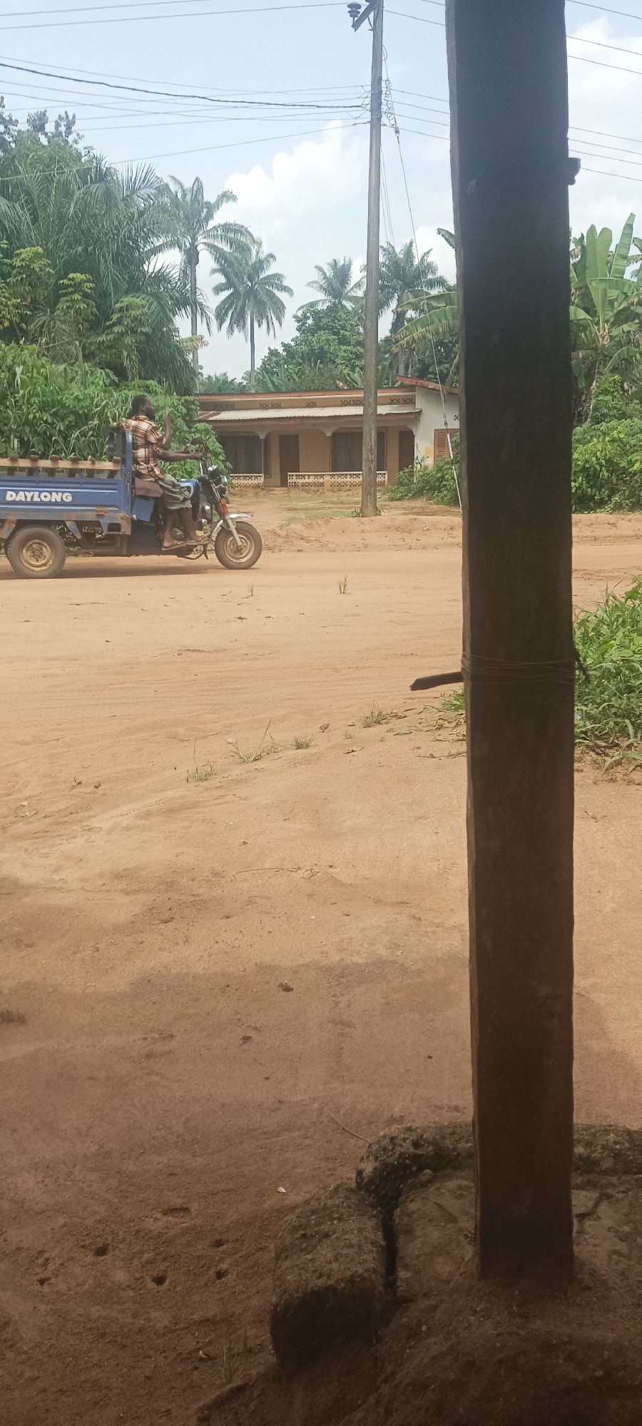 The image depicts a dirt road scene in Amauzu, Nigeria. A blue "DAYLONG" branded tricycle is traveling along the road, carrying at least one male passenger in the back. The driver appears to be raising his hand. In the background is a single-story house with a light-colored facade and a brown roof. Behind the house are several palm trees and other tropical vegetation. An electrical pole with wires is visible near the house. In the foreground, a thick wooden post is partially blocking the view, with a wire wrapped around it. The ground surrounding the post is covered in dirt and some rocks.