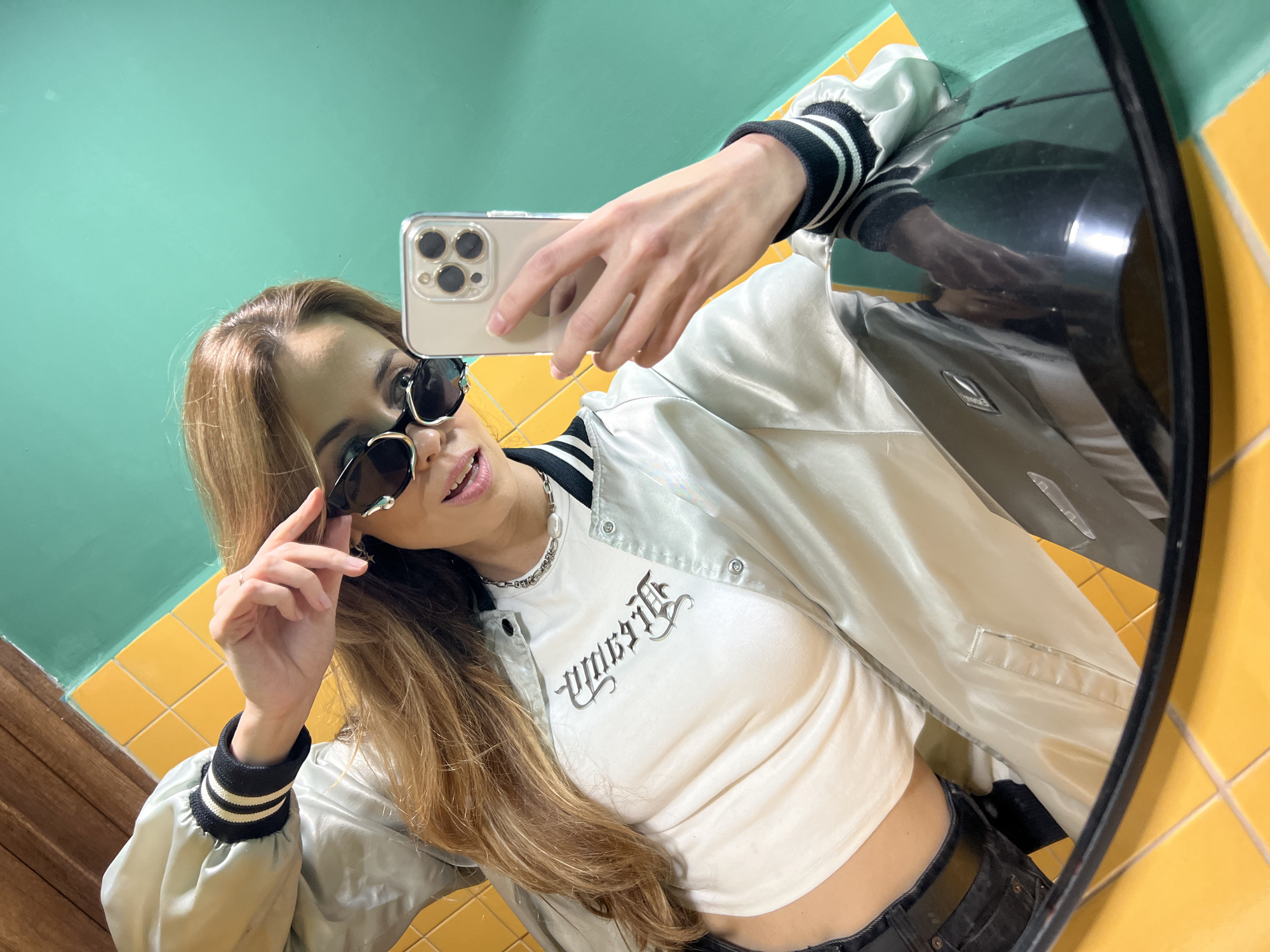 This image depicts a mirror selfie taken by a woman in a location identified as Bar, Montenegro. The woman, with long brown hair, is positioned in the foreground, leaning slightly. She is wearing a white crop top featuring black gothic-style text, a light grey or silver satin bomber jacket with black and white striped ribbed cuffs, and black pants secured with a black belt. A silver chain necklace with a single white pearl is visible around her neck. She holds a champagne-colored smartphone, identified as an iPhone with a clear case and a four-lens camera array, in her right hand to capture the image. Her left hand is raised, adjusting or holding the black-framed sunglasses she is wearing. The background consists of a bright yellow tiled surface and a prominent teal wall, with a wooden surface visible in the upper left corner. A reflective black object, possibly a counter or sink fixture, is present in the lower foreground.
