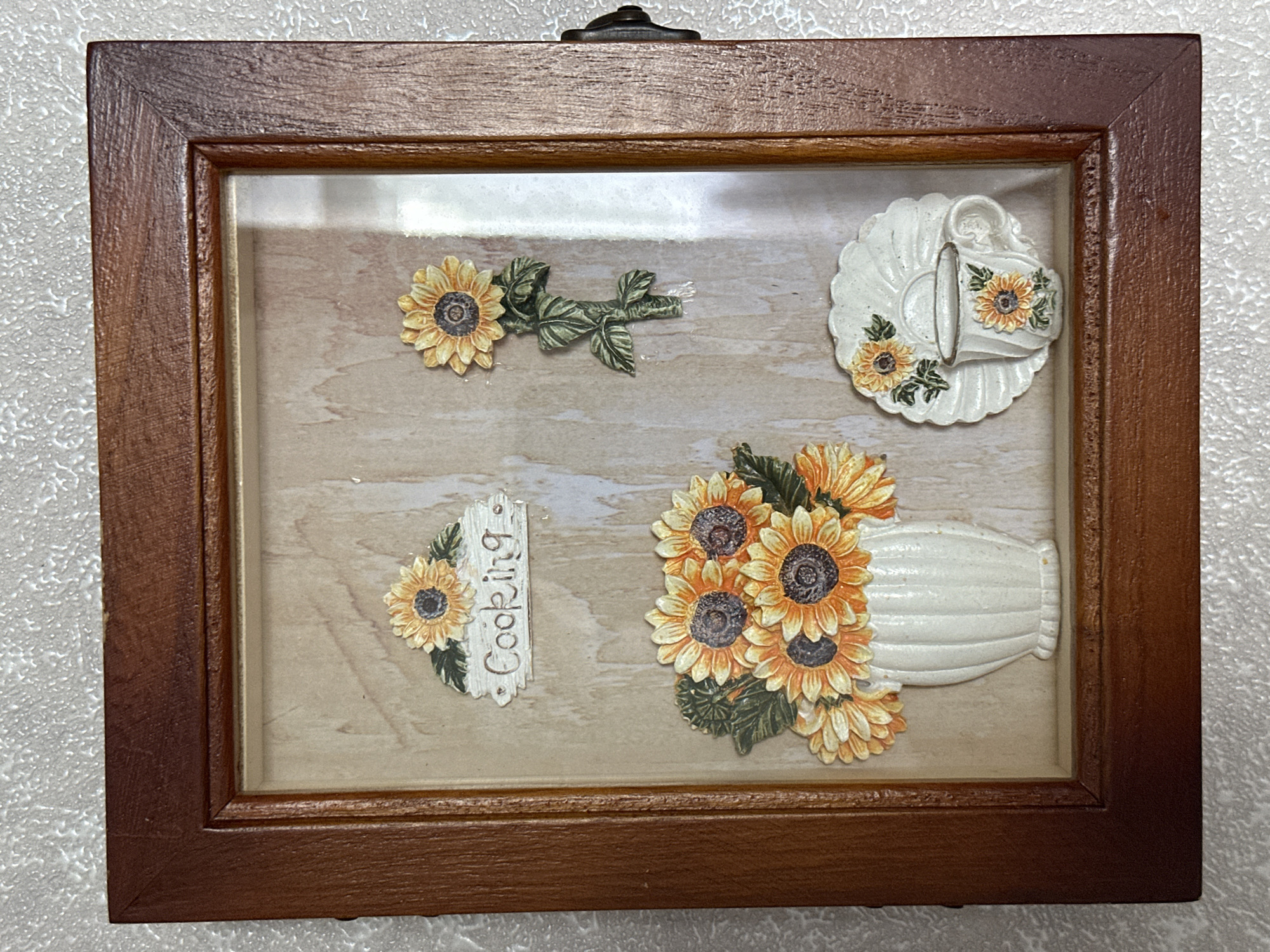 The image shows a framed decorative wall hanging with a sunflower theme. The frame is made of dark wood. Inside the frame, there is a light-colored background with a wood-grain texture. The decorative elements include a vase of sunflowers, a single sunflower on a stem, a decorative sign that says "Cooking" with two small sunflowers, and a small teacup and saucer with sunflowers. The wall hanging is likely an indoor decoration. The city of Energetik, Russia is not depicted in the image.