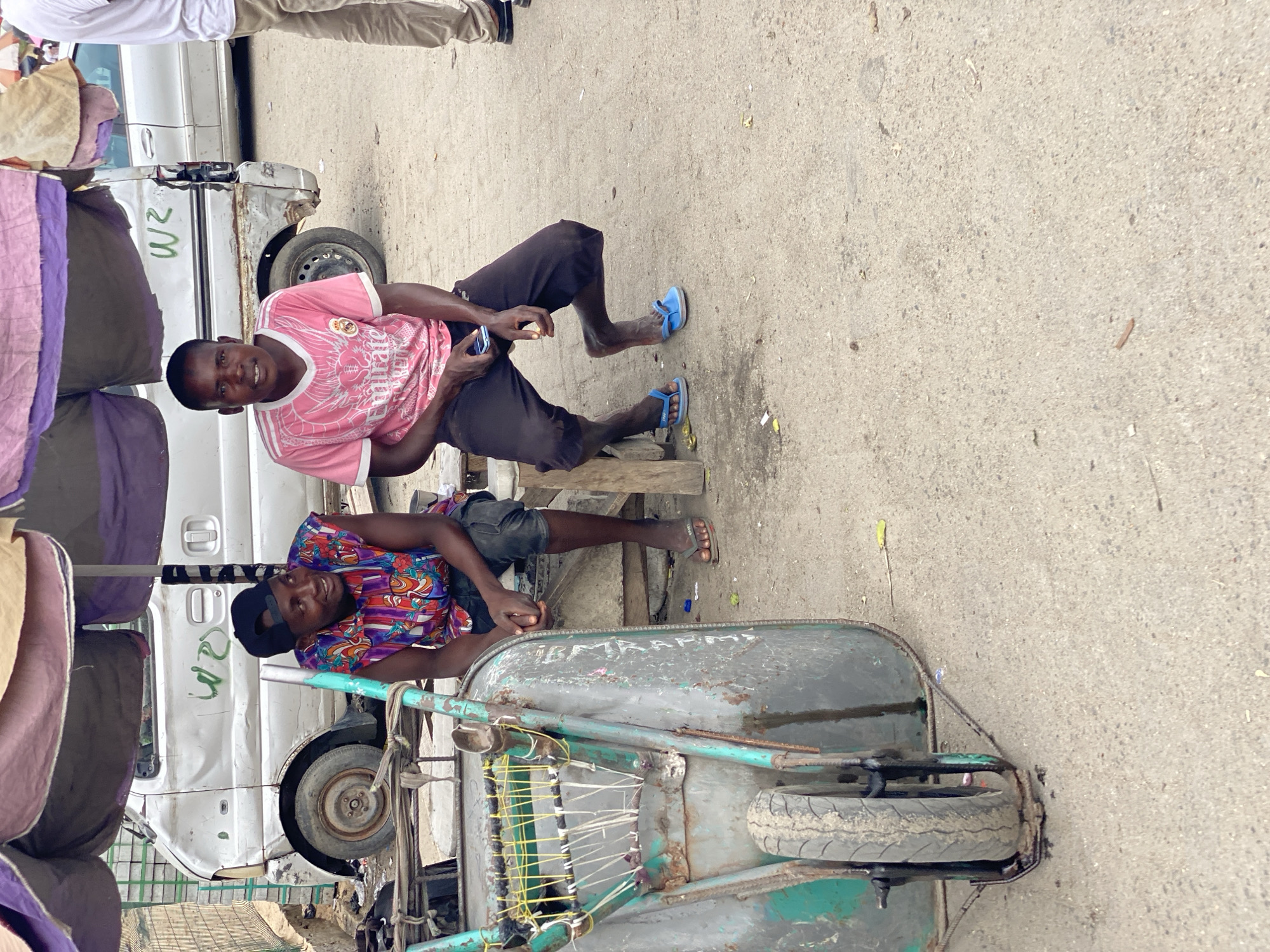 In Lagos, Nigeria, two men are seated amidst a bustling urban environment. One man, clad in a pink t-shirt and dark trousers, sits on a wooden block, casually using a mobile phone. He wears blue sandals and looks directly towards the camera with a slight smile. Adjacent to him, another man rests on a weathered wheelbarrow, wearing a patterned shirt and a dark cap, also gazing at the camera. The wheelbarrow itself is worn, with a flat tire and visible repairs. Behind them stands a dilapidated white van, marked with faded green lettering, possibly indicating its use for commercial purposes. The foreground and background are a mix of street debris and structures, suggesting a lively, possibly market-centric, urban scene. The surface is a concrete or paved road.