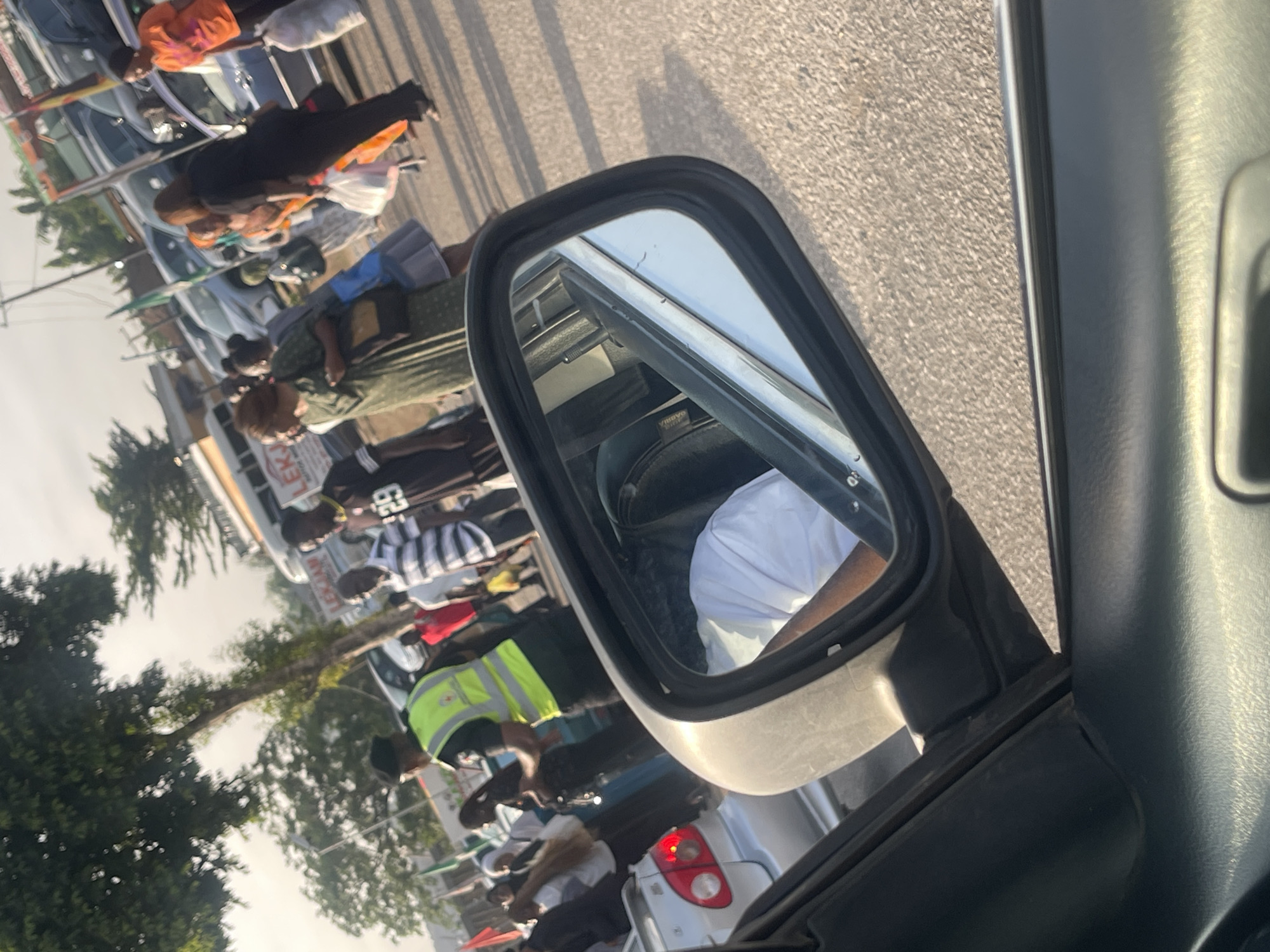 The image shows a street scene in Dutsen Alhaji, Nigeria, viewed through a car's side mirror. Several people are visible on the left side of the mirror, including individuals wearing casual clothing, a green high-visibility vest, and one in a striped shirt. A protest is unfolding. Cars are aligned on the street. The environment includes trees and buildings.