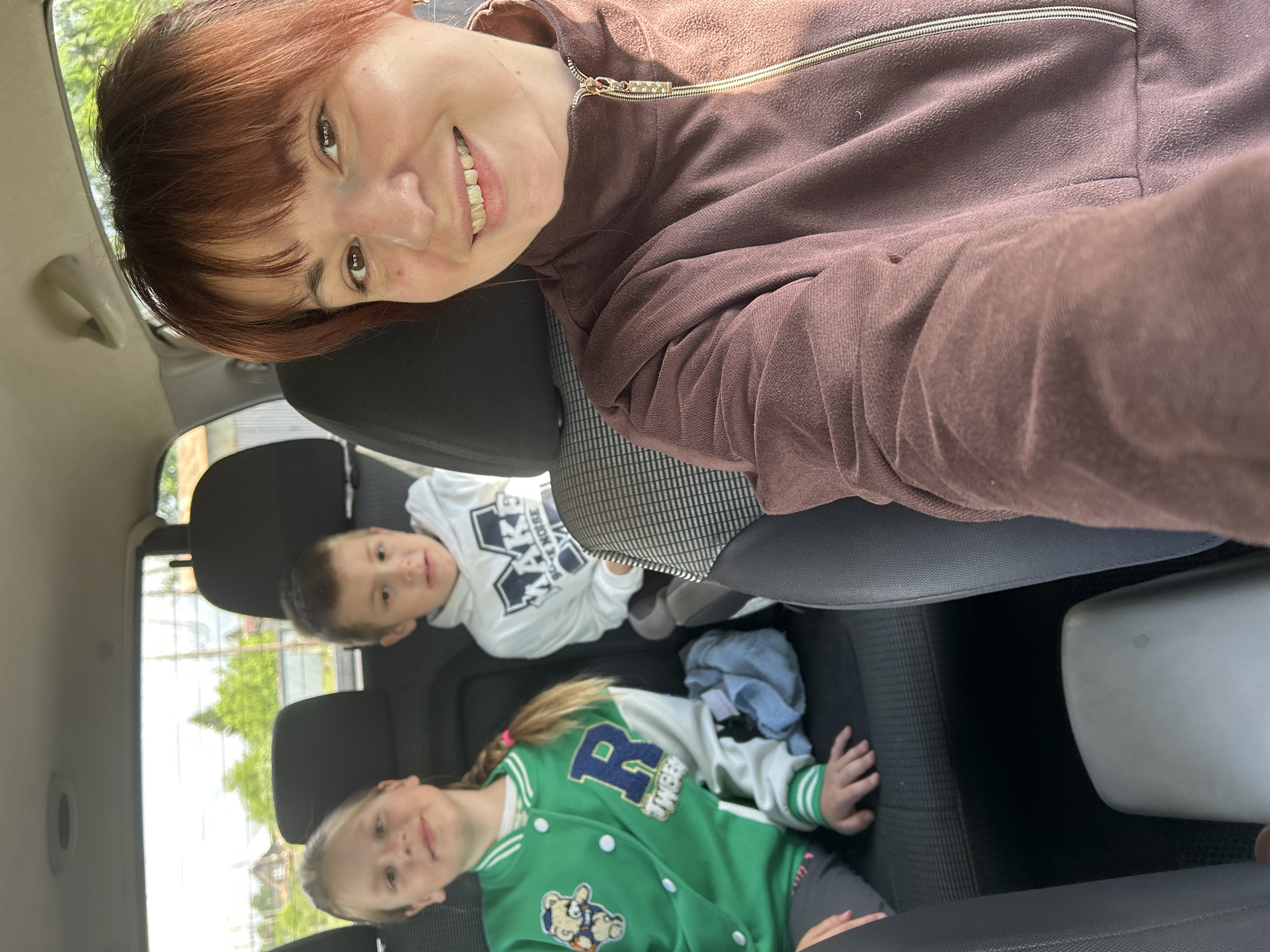 A woman is taking a selfie in the front seat of a vehicle. Two children are in the back seat. The child on the left is wearing a green jacket with a large blue "R" and a teddy bear patch. The child on the right is wearing a white hoodie with dark print. The location is Фоки, Russia.