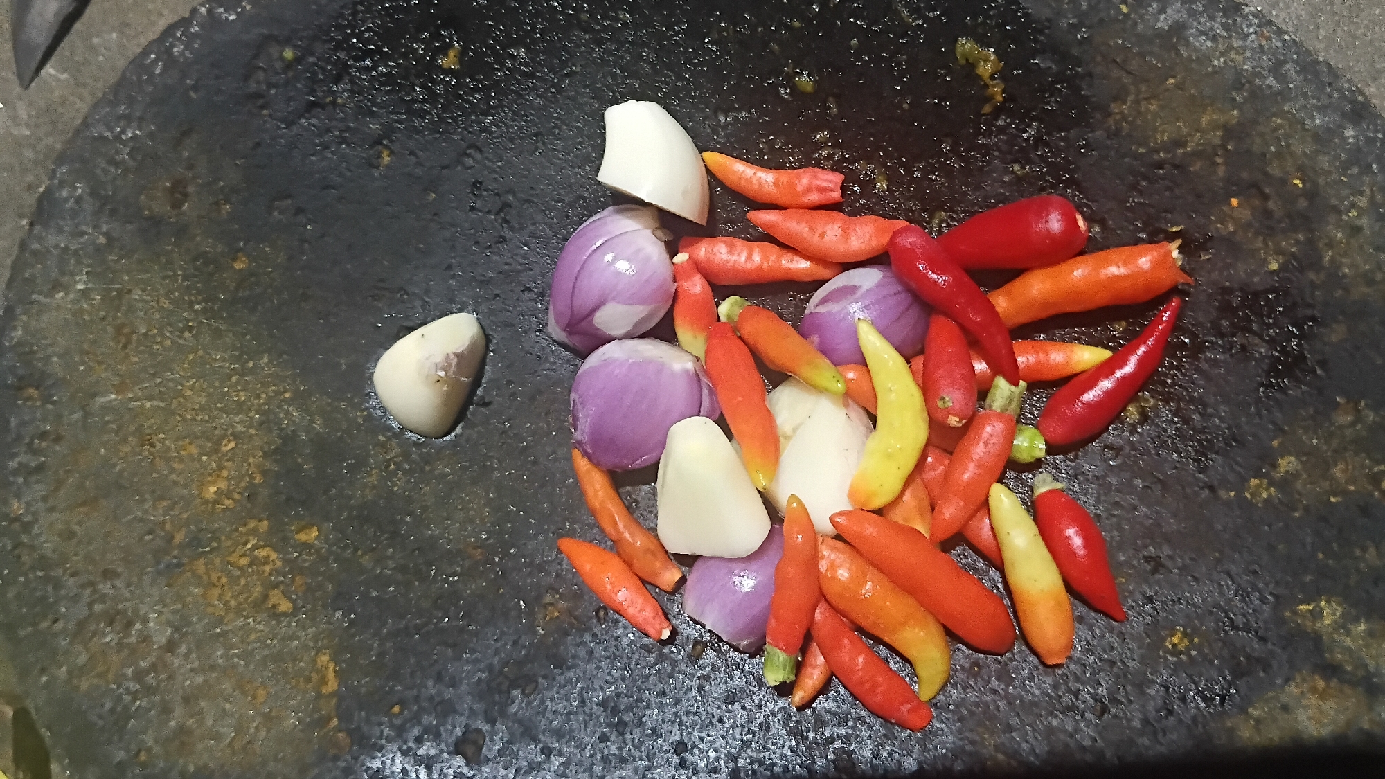 A close-up shot shows various ingredients for a sambal, a type of Indonesian chili sauce, in a dark, rough-textured mortar, likely made of stone. The ingredients include red and yellow bird's eye chilies, sliced red onions, and pieces of peeled garlic. The mortar is wet, suggesting the ingredients may have been washed or are about to be pounded. The lighting is dim, creating a moody atmosphere, and the background is out of focus. The scene is likely from a kitchen in Prabumulih, Indonesia, where sambal is a common condiment.
