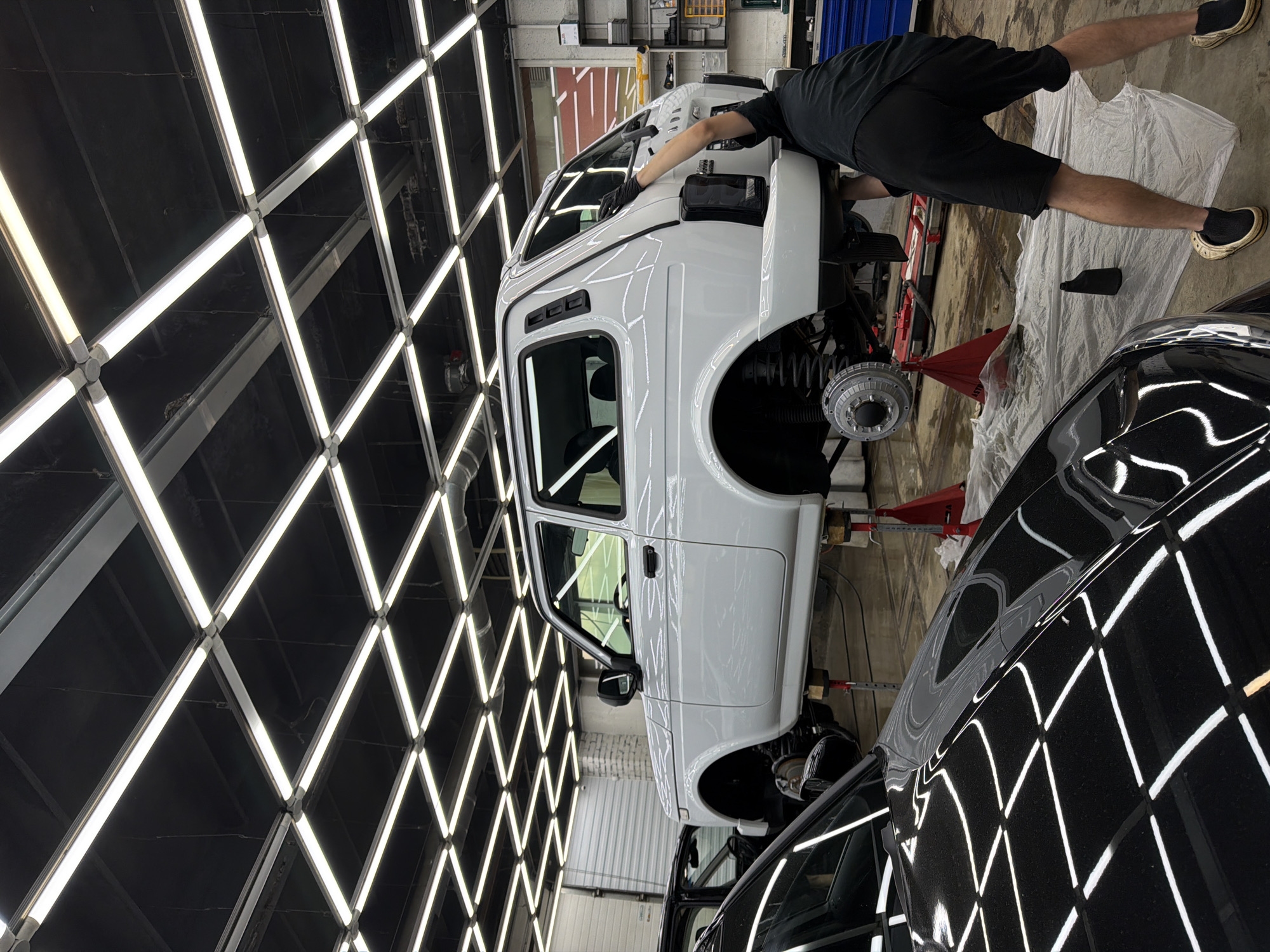 In an indoor automotive workshop in Krasnoyarsk, Russia, a white Lada Niva is elevated on red hydraulic lifts, undergoing service. A person, dressed in black shorts and a t-shirt, with black gloves, is seen working on the rear of the vehicle. The workshop ceiling is a grid of bright fluorescent lights. In the foreground, the polished black hood of another vehicle reflects the overhead lights. The floor is covered with a plastic sheet.