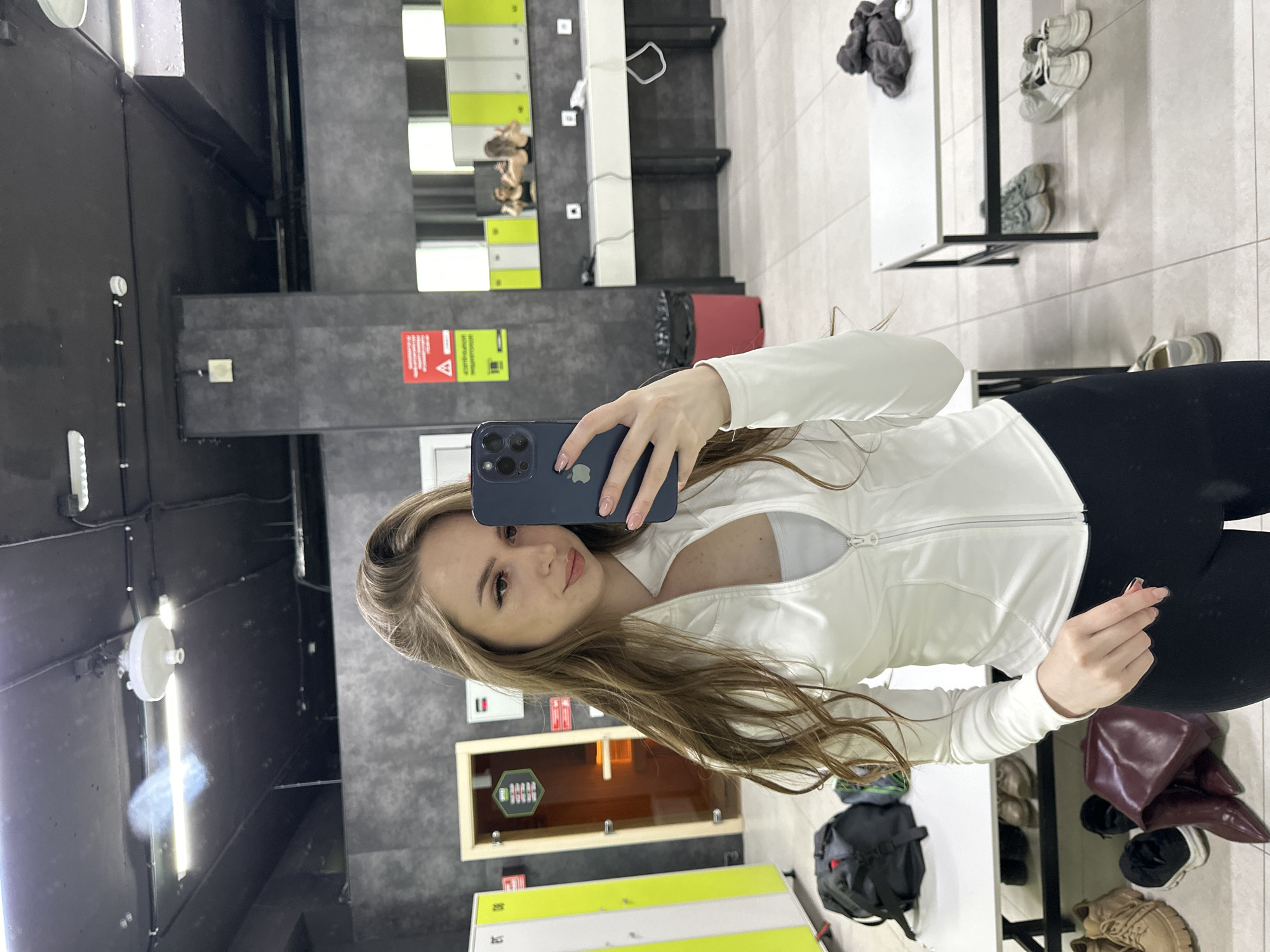 A woman takes a selfie in what appears to be a locker room. The room has grey walls and flooring. Lockers are visible in the background. A mirror reflects the scene. Several items, including shoes and bags, are scattered around the floor. The woman is wearing black leggings, a white top, and holding a blue smartphone.
