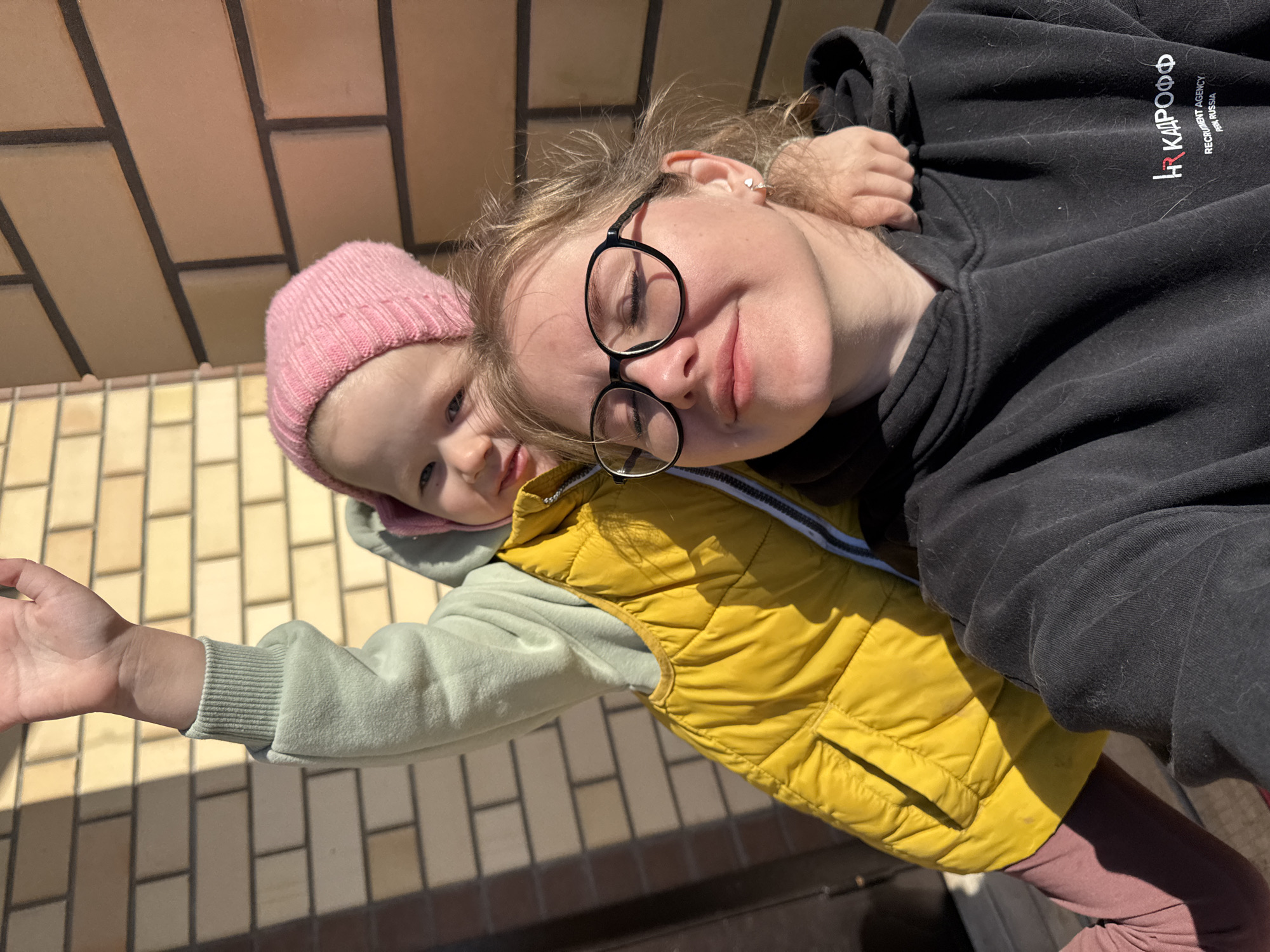An outdoor selfie captures a woman with round glasses, wearing a black hoodie with Cyrillic writing, holding a child wearing a pink knitted hat and a yellow vest over a green sweatshirt. The scene is located in Gornyi (Dvurechenskoe S/P), Russia, with a light brick surface visible in the background. The child's left arm is extended towards the camera.