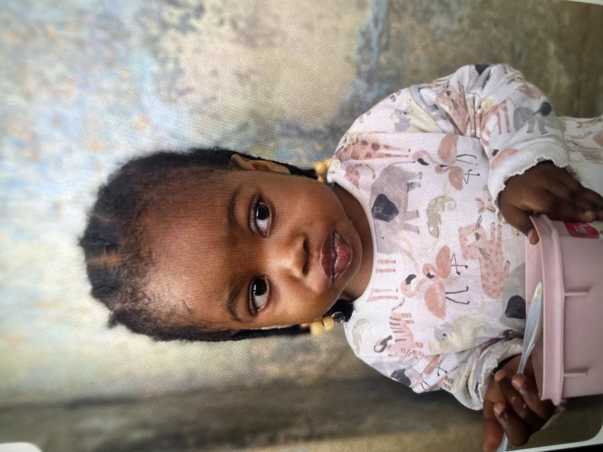 A young Black child is shown from the chest up, looking to the right of the frame. The child has braided hair with yellow beads and is wearing a long-sleeved shirt with a pattern of animals including flamingos, elephants, and giraffes. The child is holding a white spoon in their right hand, which is partially inside a light pink bowl. The background is out of focus and appears to be a textured wall. The setting is unknown.